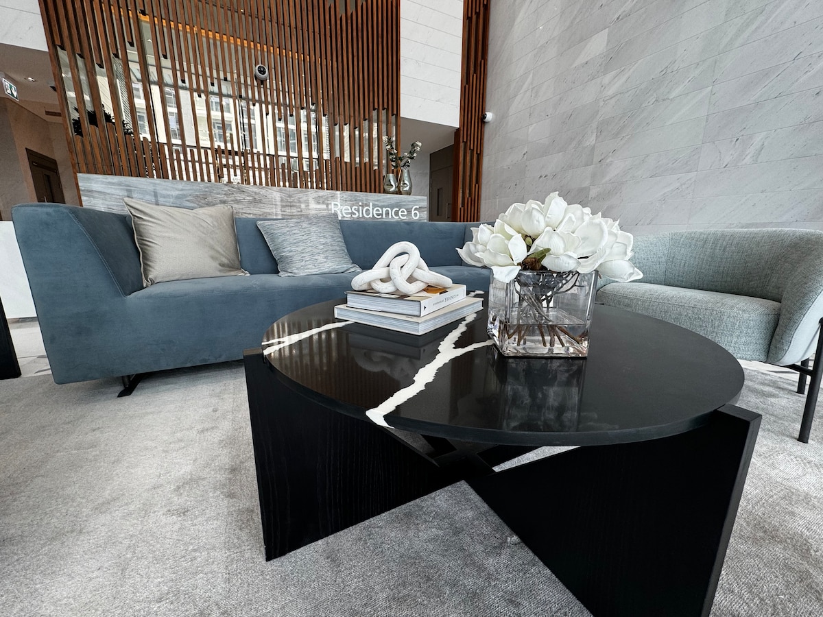A modern living area is presented with a plush gray sofa adorned with decorative cushions. A sleek black table with a marble inlay is positioned in front, holding a glass vase with white flowers and neatly stacked books, enhancing the space's contemporary design.