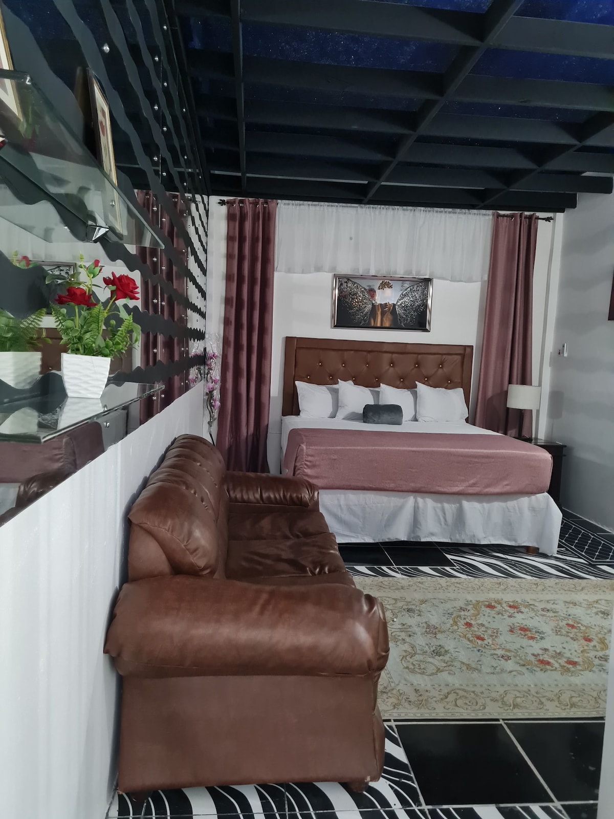 A spacious room is showcased, featuring a large bed with a tufted headboard. A cozy brown sofa is positioned nearby, accompanied by a decorative rug. Bright flowers in a vase add a touch of color, while a piece of art hangs above the bed, contributing to the overall inviting ambiance.