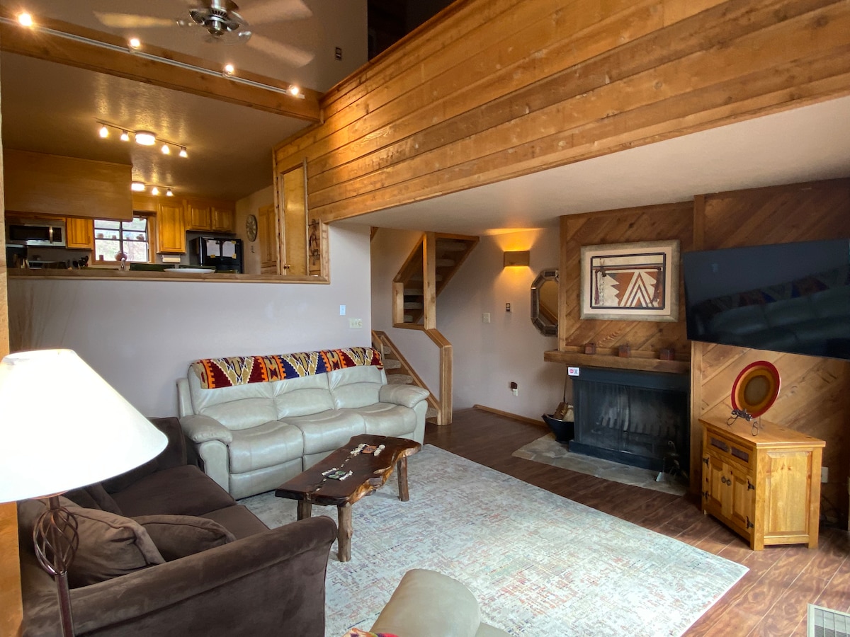 A spacious living area features comfortable seating arrangements with a light-colored couch and a dark sofa. A wooden coffee table is centered on a patterned rug. The room is illuminated by ceiling lights and includes a staircase leading to the upper level.