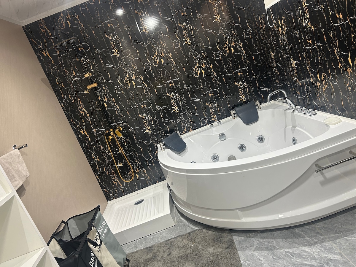 A modern bathroom features a spacious whirlpool tub with multiple jets, set against a stylish black and gold marbled wall. A shower area with a sleek floor is visible, and the room is accented with a neutral-toned rug and organized storage solutions.
