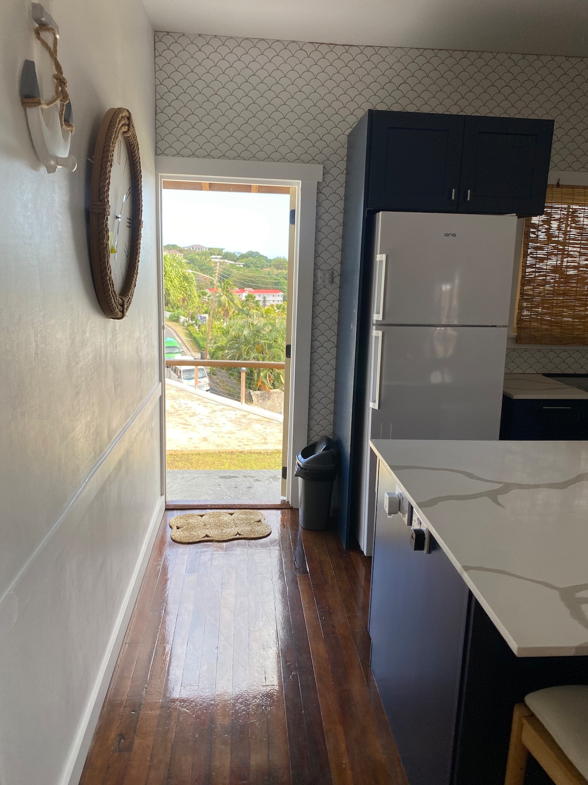 A modern kitchen is featured with dark cabinetry and a sleek countertop. A doorway leads outside, providing natural light and a view of the surroundings. A textured wall adds visual interest, while the warm wooden floor contributes to a cozy ambiance.