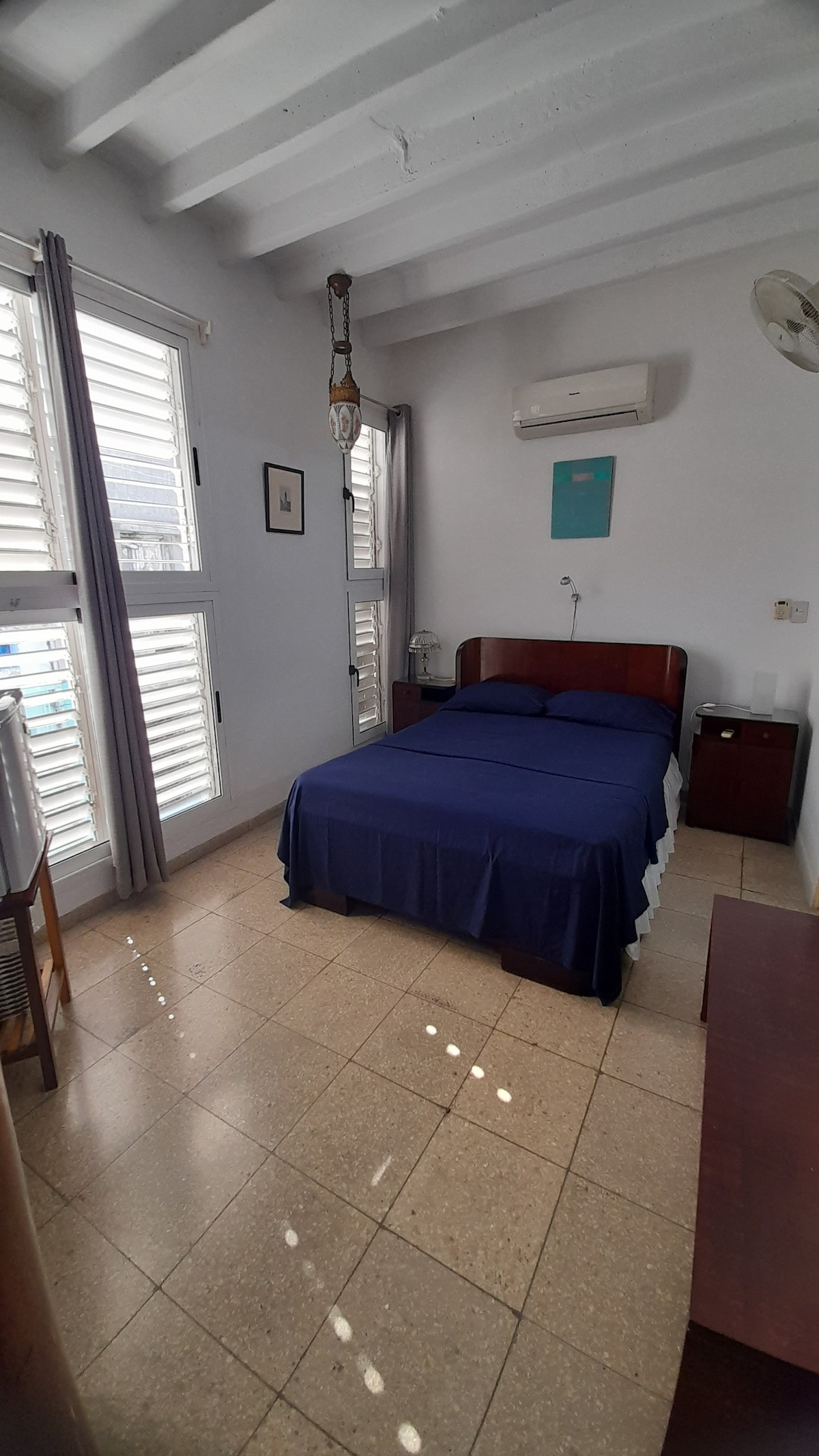 A sleeping area is arranged with a cozy bed draped in navy blue linens. Natural light filters through partially closed shutters, illuminating the neutral-toned floor. Air conditioning is mounted on the wall, and a bedside table sits alongside the bed.