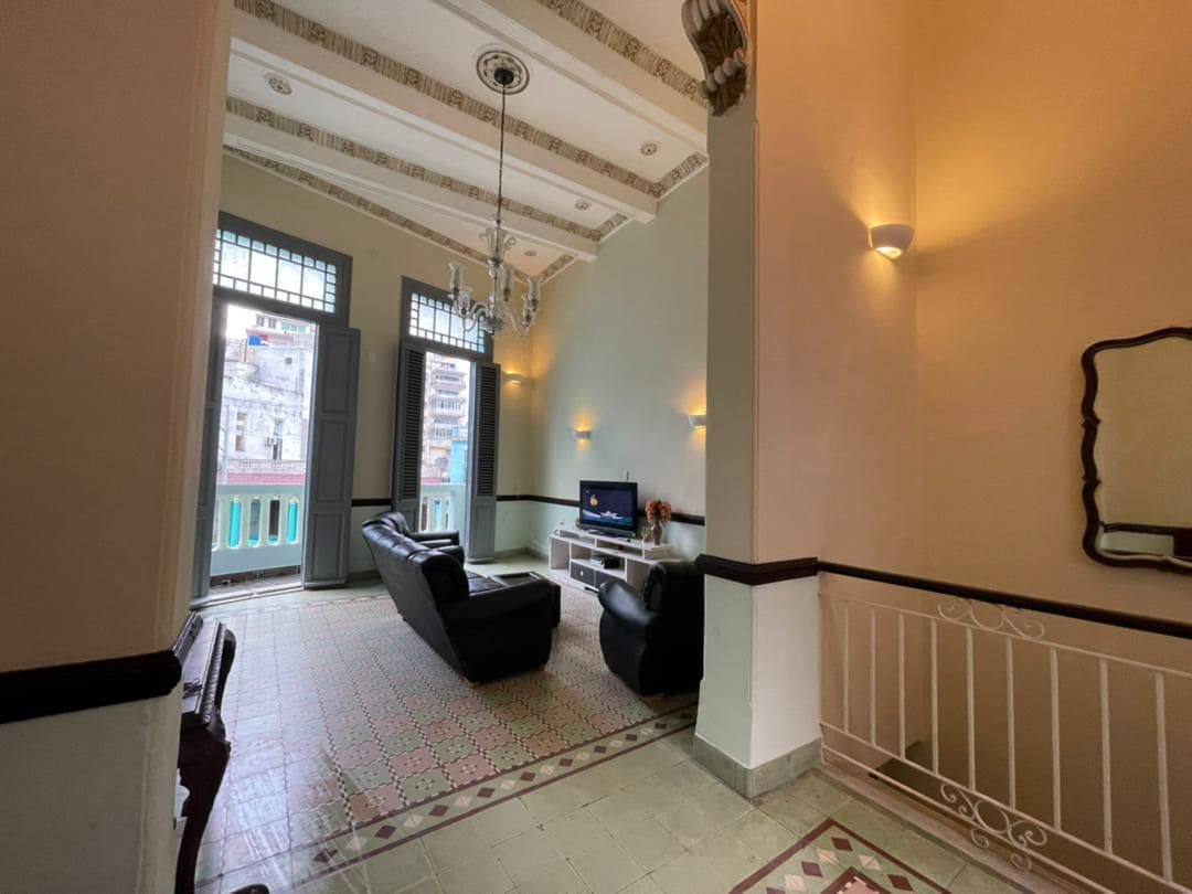 The living room features high ceilings with decorative molding and ample natural light from large windows. Two black sofas are positioned in front of a modern television, and a vintage mirror hangs on the wall. The distinctive tiled flooring adds character to the space.