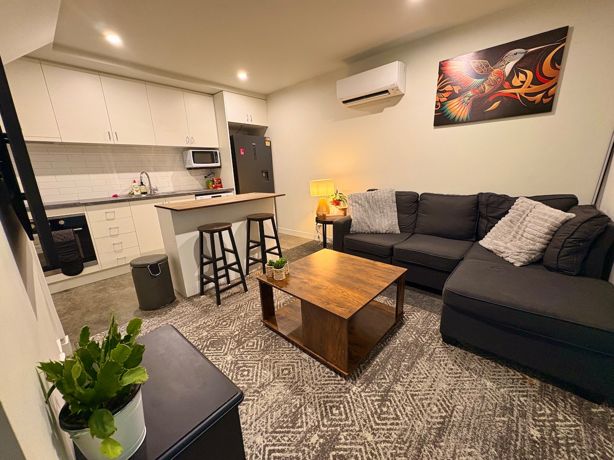 The living area features a cozy sectional sofa, a wooden coffee table, and a decorative rug. A contemporary kitchen with white cabinetry is visible, equipped with an oven, microwave, and seating bar. A piece of wall art adds character, while plants enhance the space.