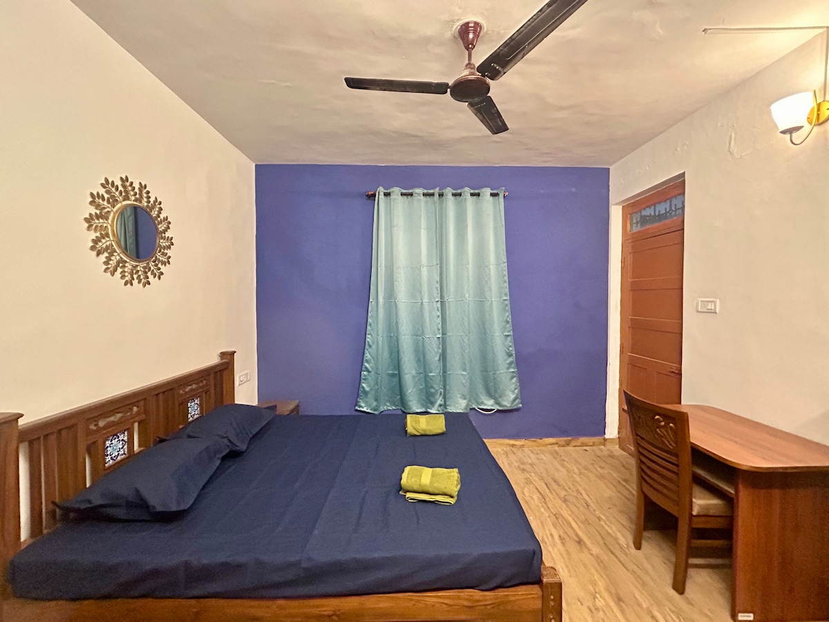A spacious room is presented with a dark blue accent wall, featuring a wooden bed adorned with navy bedding. Two green towels are neatly placed on the bed. A study table and chair occupy one corner, complemented by a ceiling fan and light fixture.
