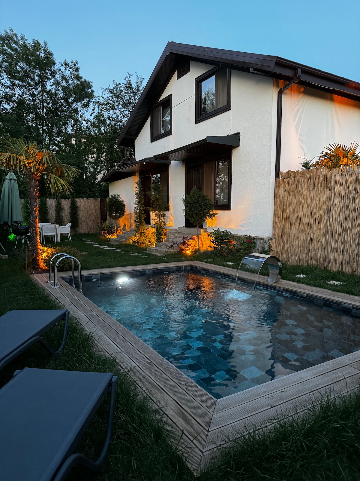 The exterior of a twin villa bungalow is visible, featuring a cozy garden area and warmly lit landscaping. A small, inviting pool is situated beside a wooden deck, complemented by lounge chairs. The house showcases large windows surrounded by greenery and a bamboo fence, enhancing privacy.