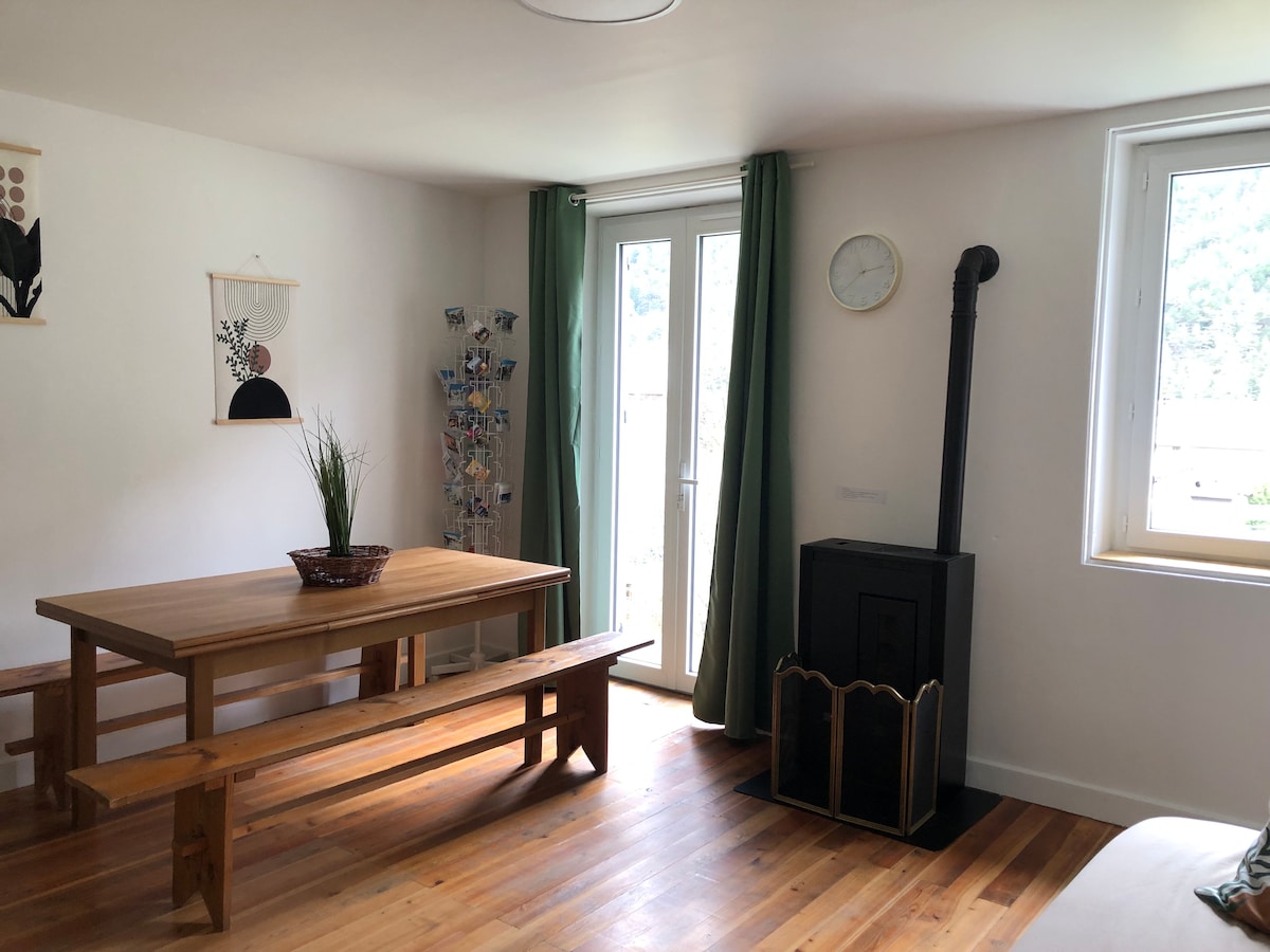 A wooden dining table with benches is positioned in the center of the inviting space. A small stove is located in the corner next to a window, framed by green curtains. Light hardwood flooring complements the neutral color scheme and natural elements.