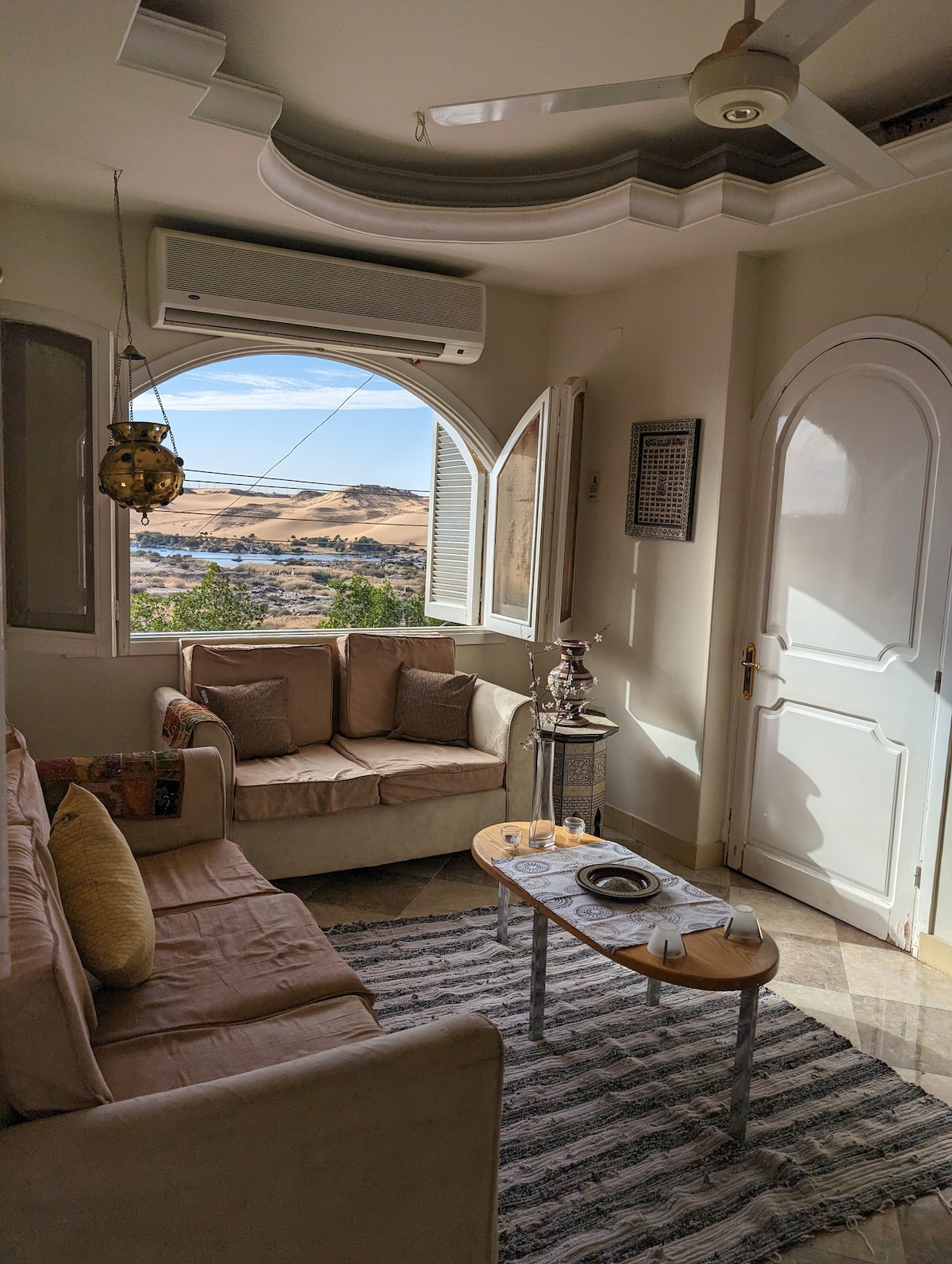 A cozy sitting area features light-colored seating arranged around a small table. Large arched windows provide views of the surrounding landscape, while decorative elements add a touch of character. Air conditioning ensures comfort in the space, which is brightened by natural light.