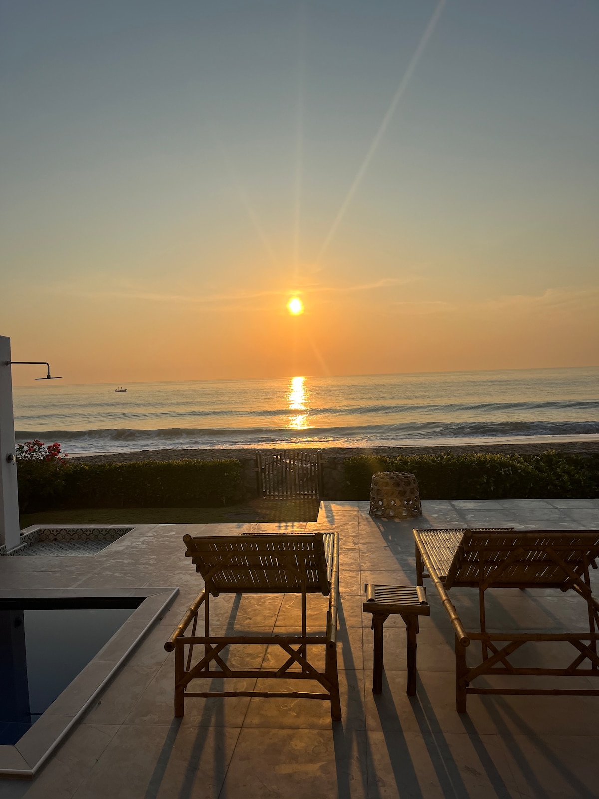 A serene view of the sunset over the ocean is captured, with sunlight reflecting on the water. Two wooden lounge chairs are positioned on the patio, facing the beach. A swimming pool is visible beside the patio, framed by lush greenery.