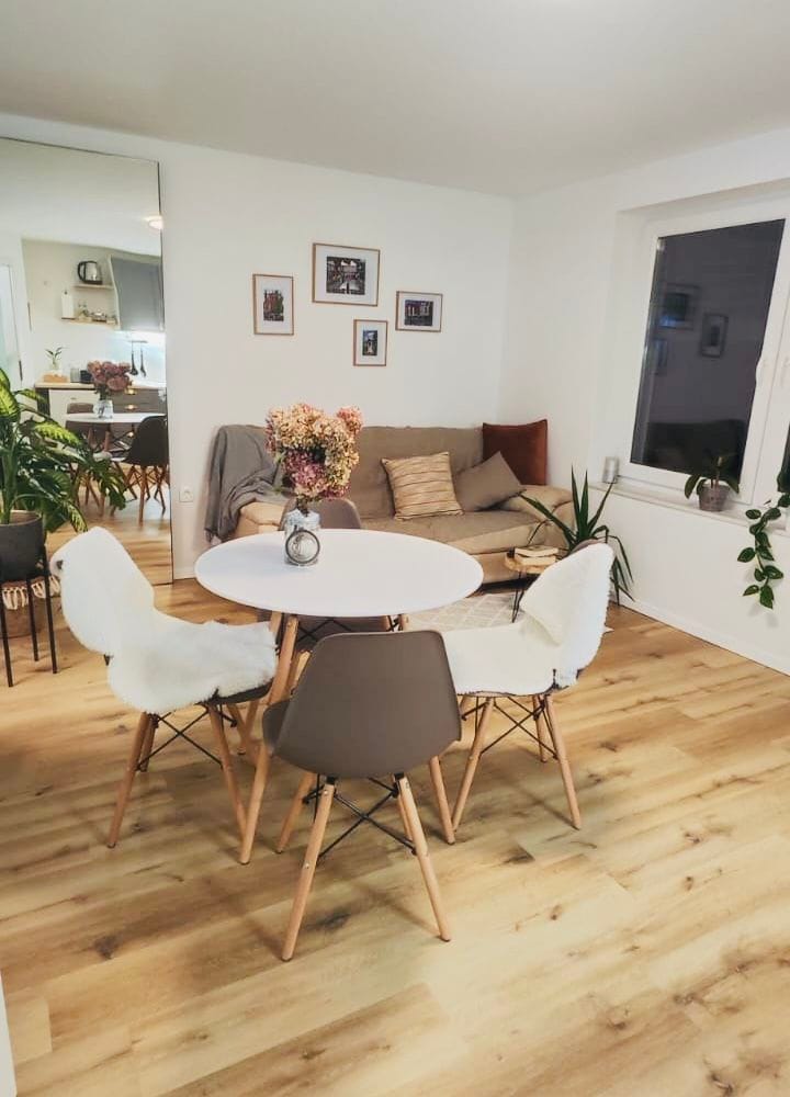 A welcoming dining area features a round table surrounded by four chairs, with one upholstered in a soft material. Fresh flowers are arranged in a small vase at the center. A cozy seating nook with cushions is visible in the background, complemented by various potted plants.