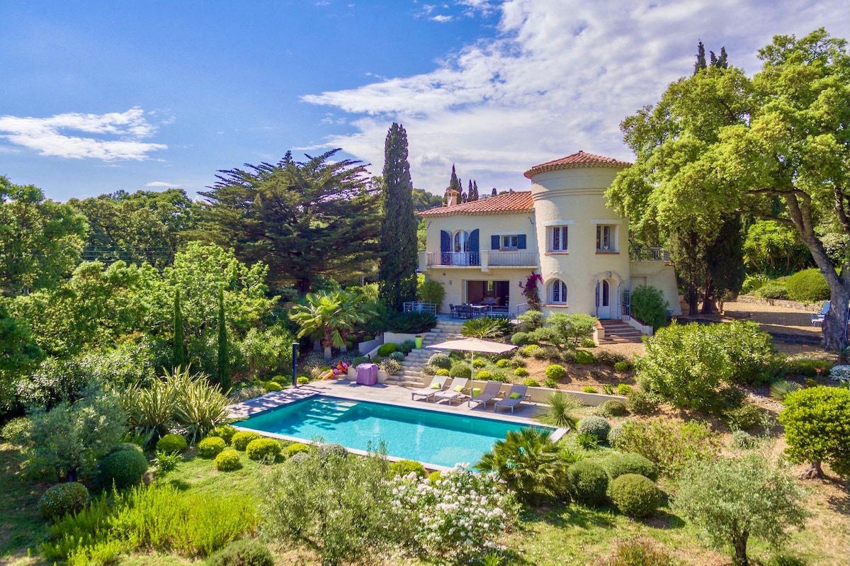 A stately villa is surrounded by a lush garden, featuring a private swimming pool with sun loungers. The charming structure is complemented by trees and shrubbery, creating a serene outdoor space. Clear skies and gentle sunlight illuminate the scene, enhancing its inviting atmosphere.