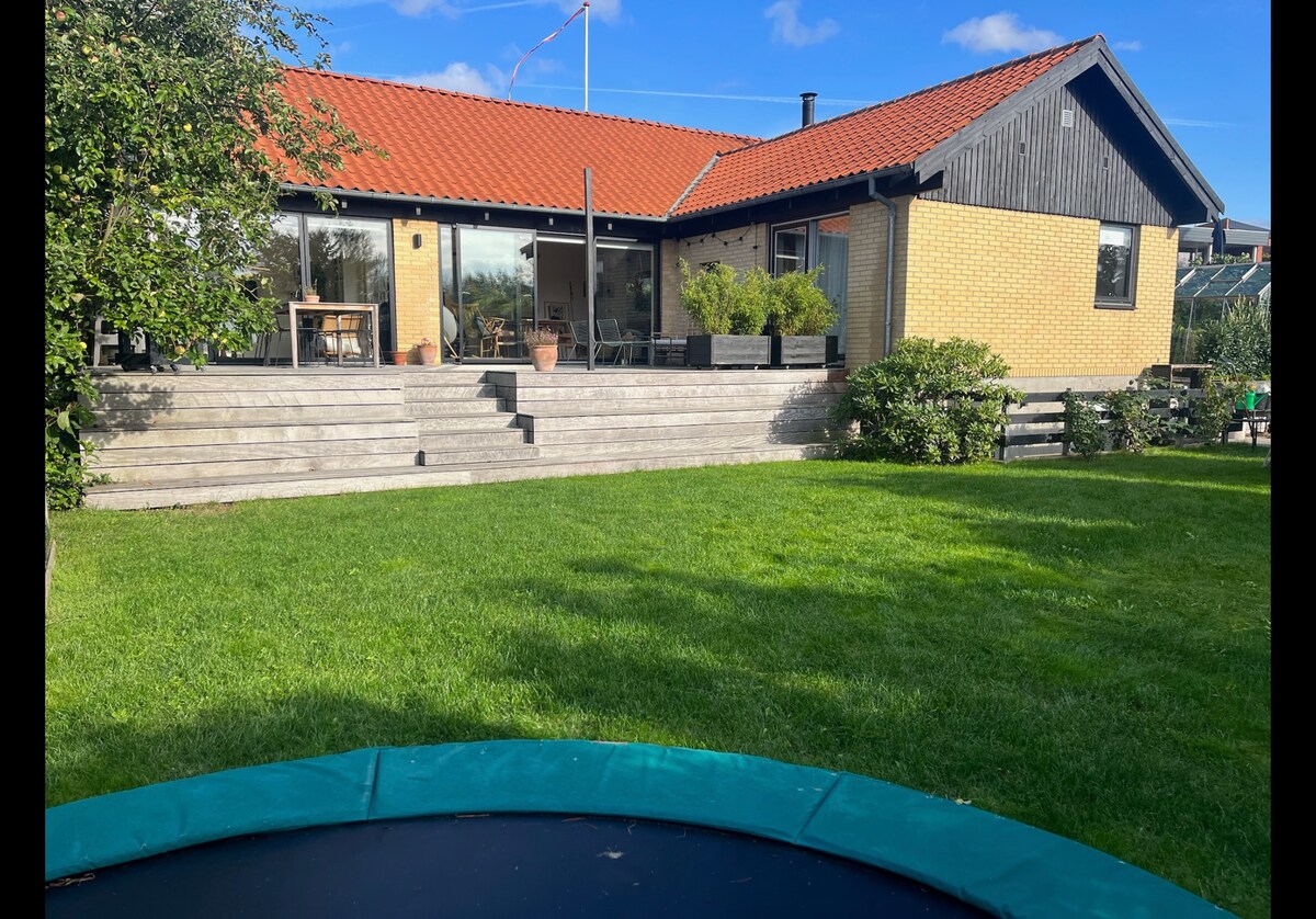 A family-friendly house is featured with a spacious lawn. The exterior showcases a blend of brick and wood, and the large deck is equipped with seating. A trampoline is visible in the green yard, inviting outdoor enjoyment. Large glass doors provide views into the home.