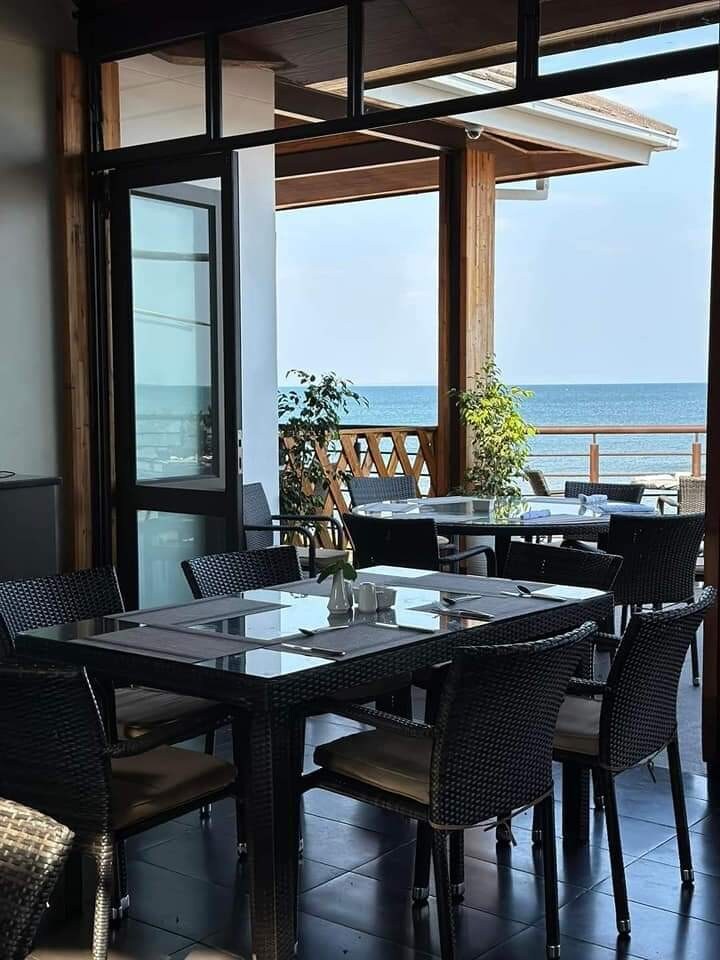 A dining area with several dark wicker chairs surrounds glass-topped tables. Large glass doors offer a view of the ocean and allow natural light to fill the space. Potted plants add a touch of greenery, enhancing the coastal ambiance.