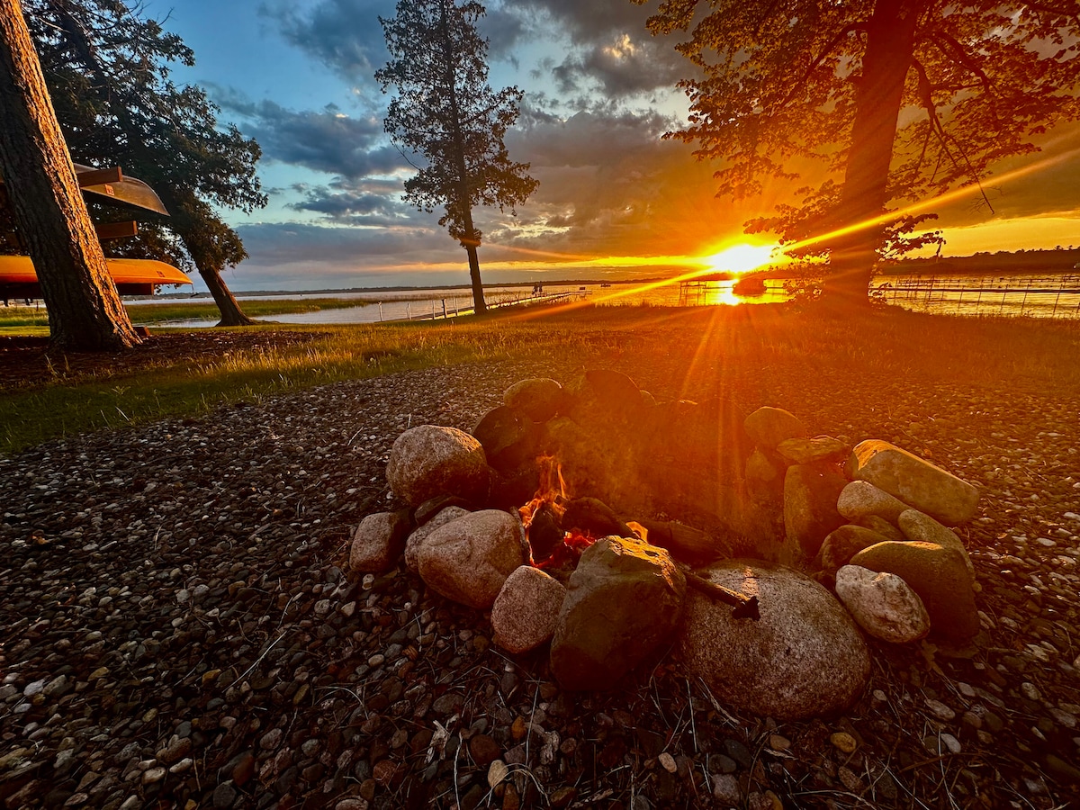 A fire pit constructed with smooth stones is centered on a pebbled area, surrounded by tall trees. The horizon showcases a setting sun, casting warm hues across the serene lake. Two canoes are visible above the water's edge, enhancing the tranquil lakeside setting.