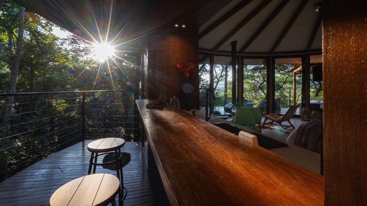 An inviting bar area features a smooth wooden counter with two stools. Sunlight streams through the open space, illuminating the surroundings. The cabin's design highlights its connection to the natural environment, with large windows offering views of the lush greenery outside.