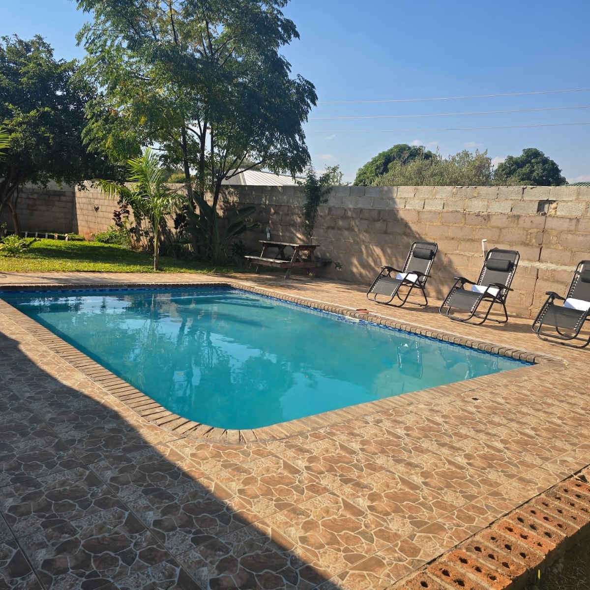 A swimming pool is surrounded by a paved patio, featuring several loungers. Lush greenery is visible in the background, along with a shaded area with picnic-style seating. The clear water reflects the blue sky, providing a refreshing getaway ideal for relaxation.