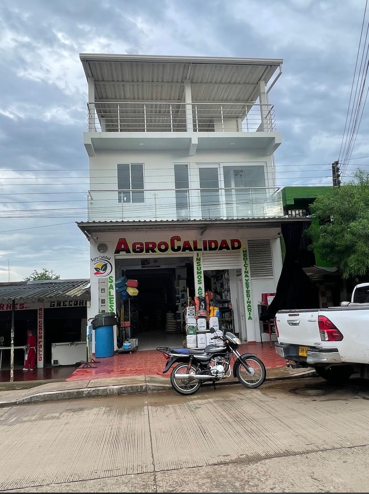 A three-story building is presented with a modern façade. The first level features a storefront with signage that reads 'AgroCalidad.' A motorcycle is parked in front, and the upper levels are equipped with several large windows, allowing natural light to fill the interior.