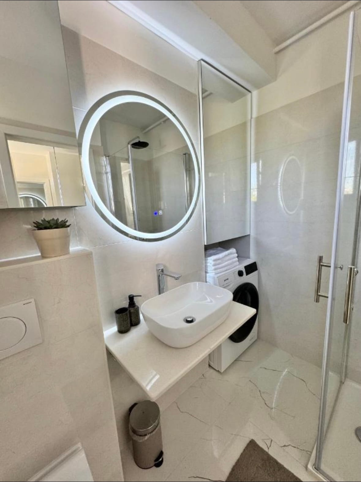 A modern bathroom features a circular mirror above a sleek sink, complemented by stylish dark fixtures. A spacious shower enclosure is visible, along with neatly arranged white towels. A washing machine is integrated into the design, and natural light fills the area through a window.