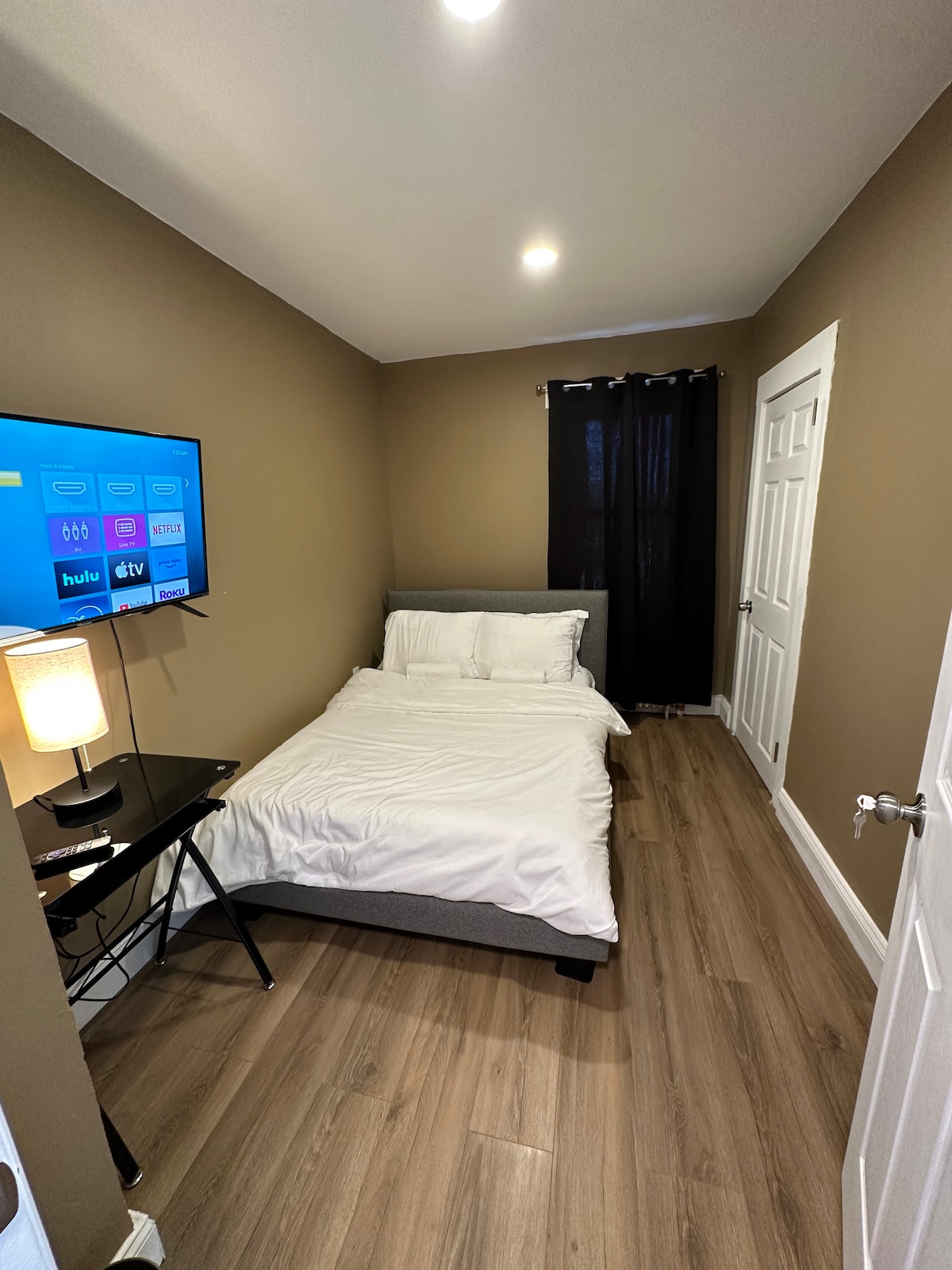 A compact bedroom is illuminated by overhead lights. A full-size bed, covered with white bedding, is positioned against the wall. A small desk holds a flat-screen TV mounted above it. Dark curtains frame a window, and a white door leads out of the room.
