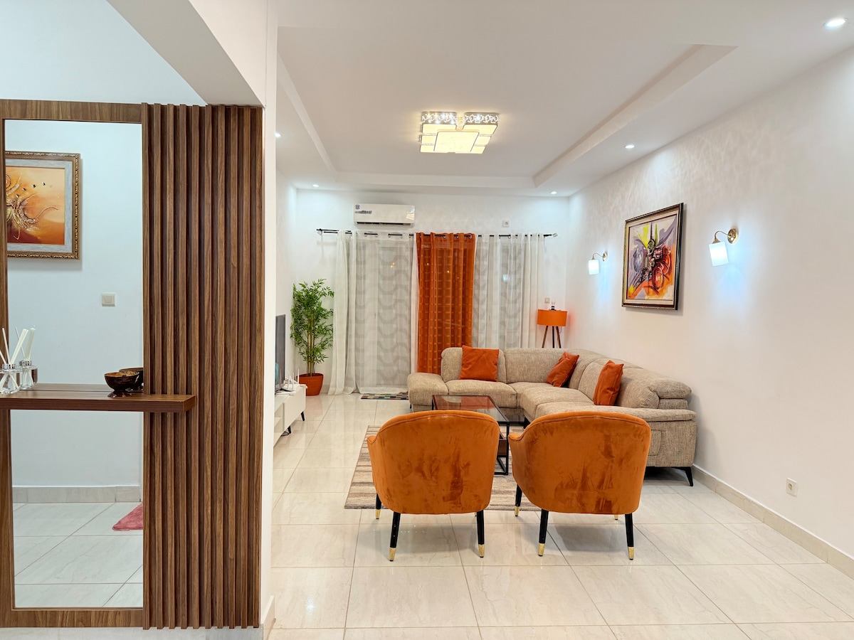 A well-lit living area is presented, featuring a spacious, L-shaped sectional sofa in muted tones with vibrant orange accent chairs. Soft lighting fixtures complement the warm color palette, while light curtains allow natural light to filter through. Decorative artwork adds character to the space.