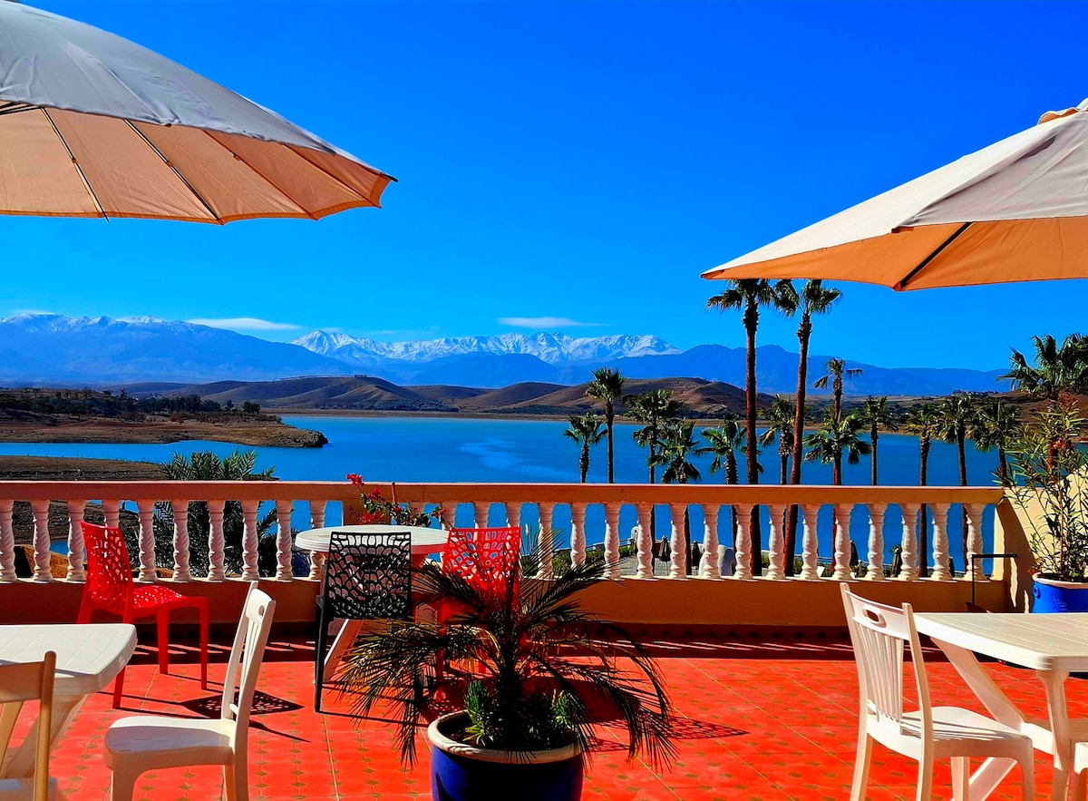 A terrace space is presented with several tables and chairs under large umbrellas, offering a view of a serene lake and distant mountains. Colorful chairs are complemented by potted plants, creating an inviting outdoor environment.
