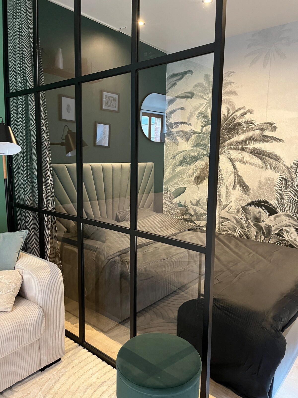 A cozy sleeping area is separated by a glass partition, featuring a plush headboard against a green wall adorned with tropical palm wallpaper. Soft lighting enhances the intimate atmosphere, and a neutral-toned sofa is positioned nearby, contributing to a stylish and functional layout.