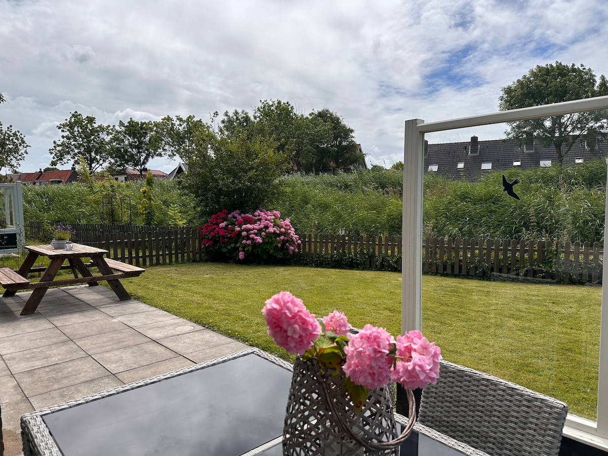 A serene outdoor space features a private garden with well-maintained grass and blooming pink flowers. A patio set with a glass tabletop is placed in the foreground, while a picnic table is situated on a tiled area. A wooden fence and lush greenery create a peaceful backdrop.