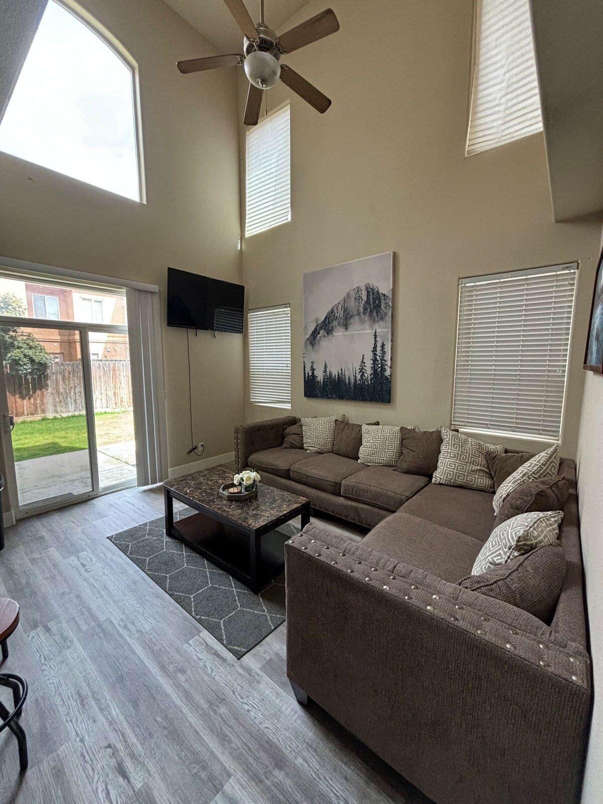 A cozy living area features a sectional sofa with decorative cushions and a wall-mounted television. Large windows allow natural light to fill the space. A sliding glass door offers access to the outdoor area, while a small coffee table sits at the center, accompanied by a decorative centerpiece.