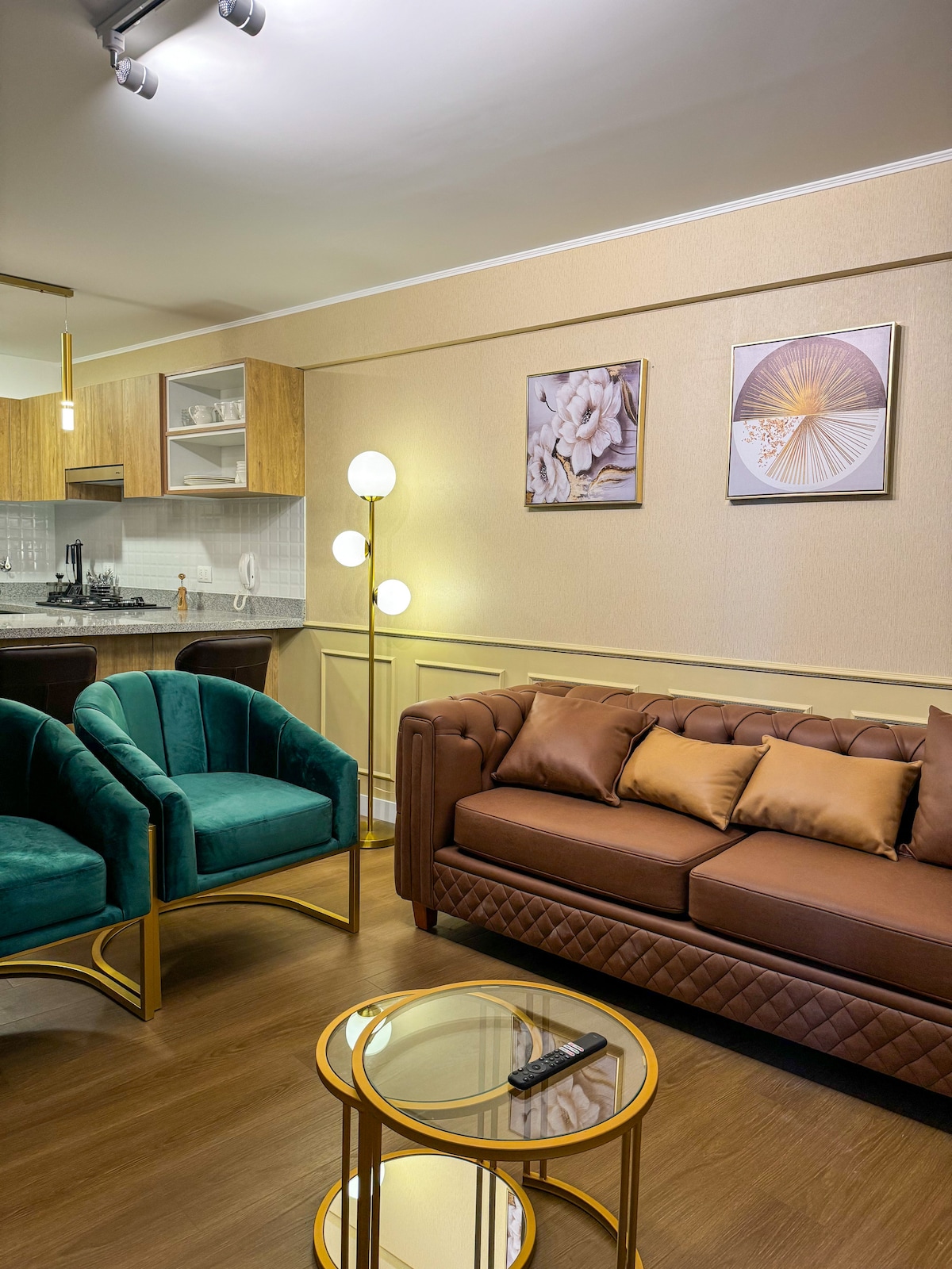 A stylish living area features a brown tufted sofa complemented by plush brown cushions. Two teal accent chairs add color, while a round golden coffee table sits in front. Behind the seating, an open kitchen is visible, accented by decorative wall art.