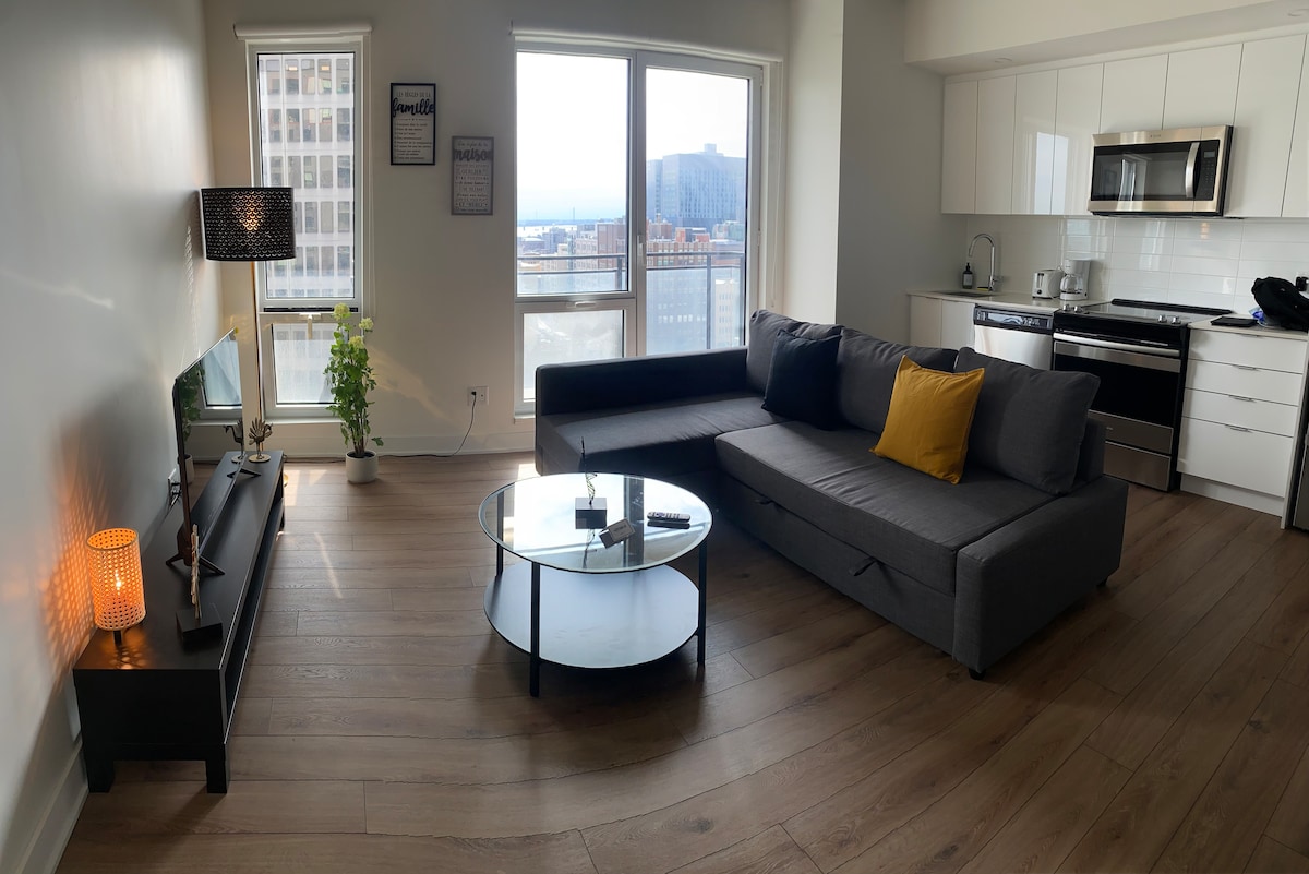 A living area features a modern sectional sofa with a yellow accent pillow, positioned near a glass coffee table. Large windows provide natural light and offer city views. A flat-screen TV is situated on a stylish console, complemented by a lamp and a well-equipped kitchenette.