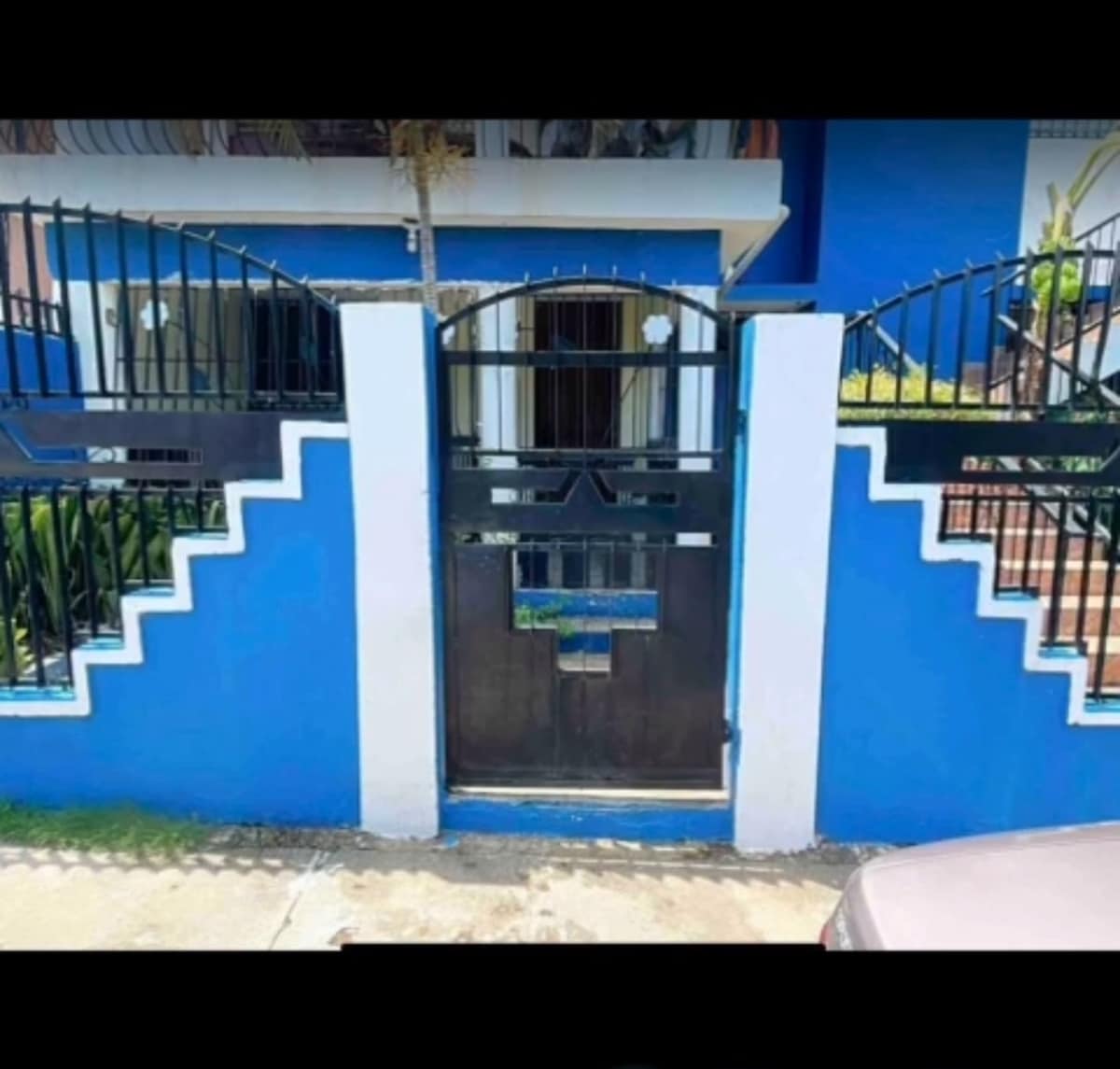 An exterior view showcases the entrance gate of the property, framed by a striking blue wall. The gate is constructed from black metal with a patterned design, bordered by minimalist white steps that lead up to the entrance.