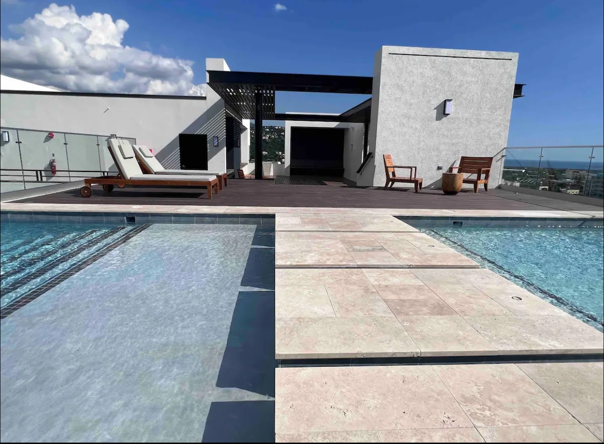 The image captures a rooftop space featuring a stunning pool with a stepped entrance, surrounded by wooden decking. Two loungers are positioned nearby, and a partial view reveals the open-air structure that offers a blend of comfort and relaxation.