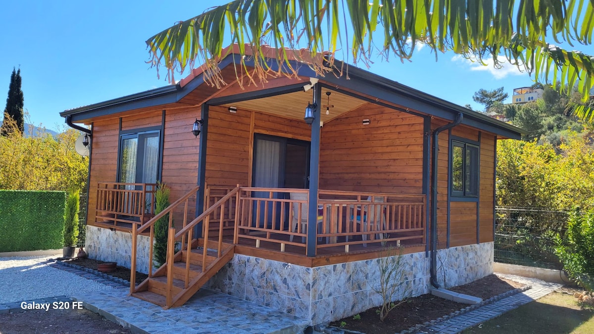 A wooden bungalow is presented with a welcoming porch featuring a railing and outdoor seating. The facade displays a natural wood finish, complemented by stone details at the base. Lush greenery surrounds the home, enhancing the tranquil setting under a clear blue sky.