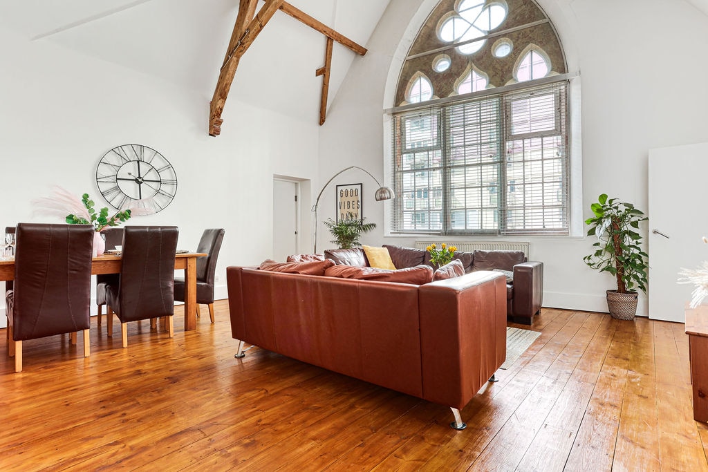 An airy living space features high ceilings with wooden beams and large windows letting in ample natural light. A comfortable leather sofa is positioned to face a wooden table, surrounded by dining chairs. A potted plant adds a touch of greenery to the warm wood floor.