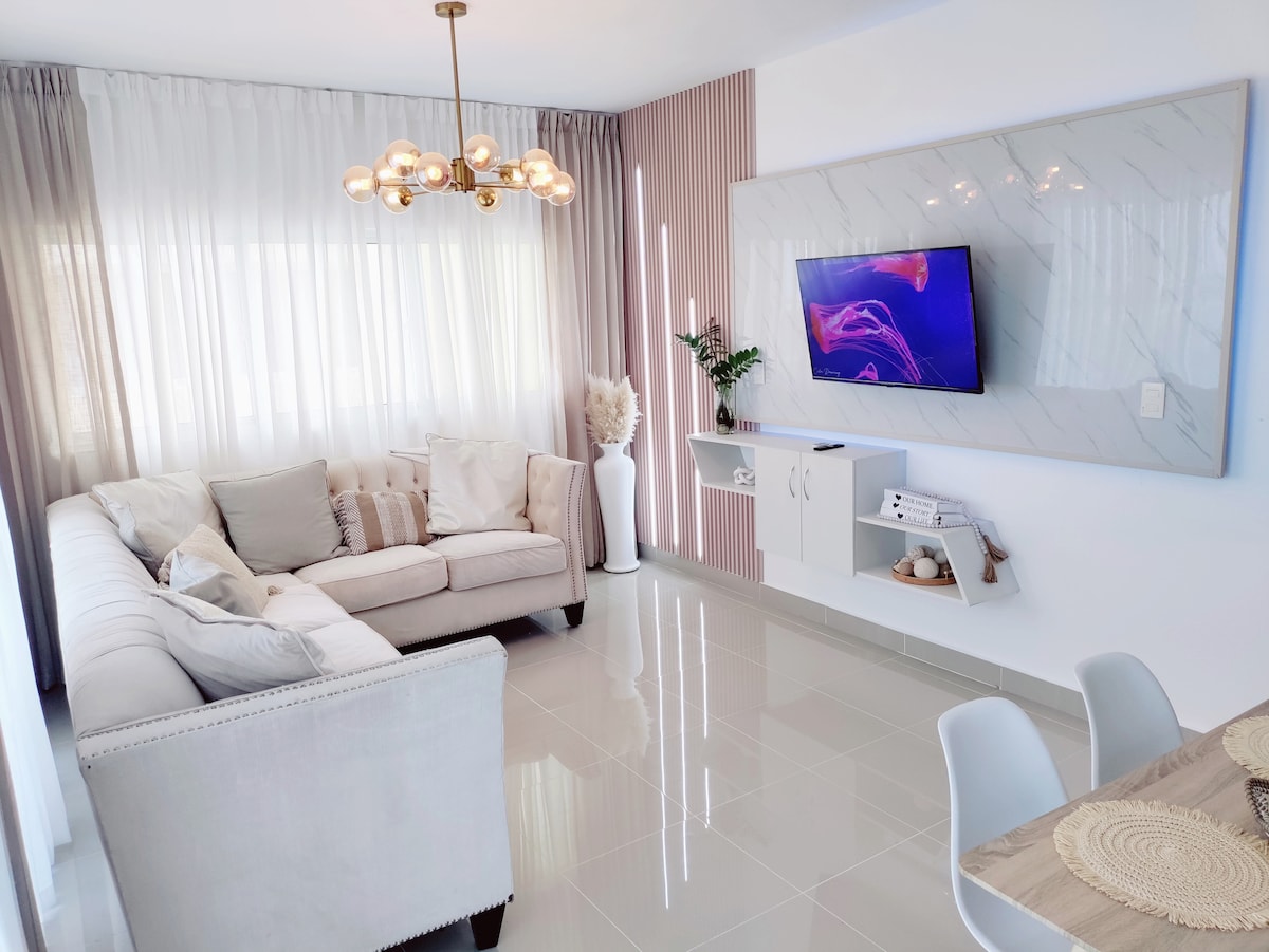 A spacious living area is illuminated by natural light filtering through sheer curtains. A comfortable sectional couch is arranged with decorative pillows. A flat-screen television is mounted on the wall above a minimalist media unit, complemented by a potted plant and sculptural elements.