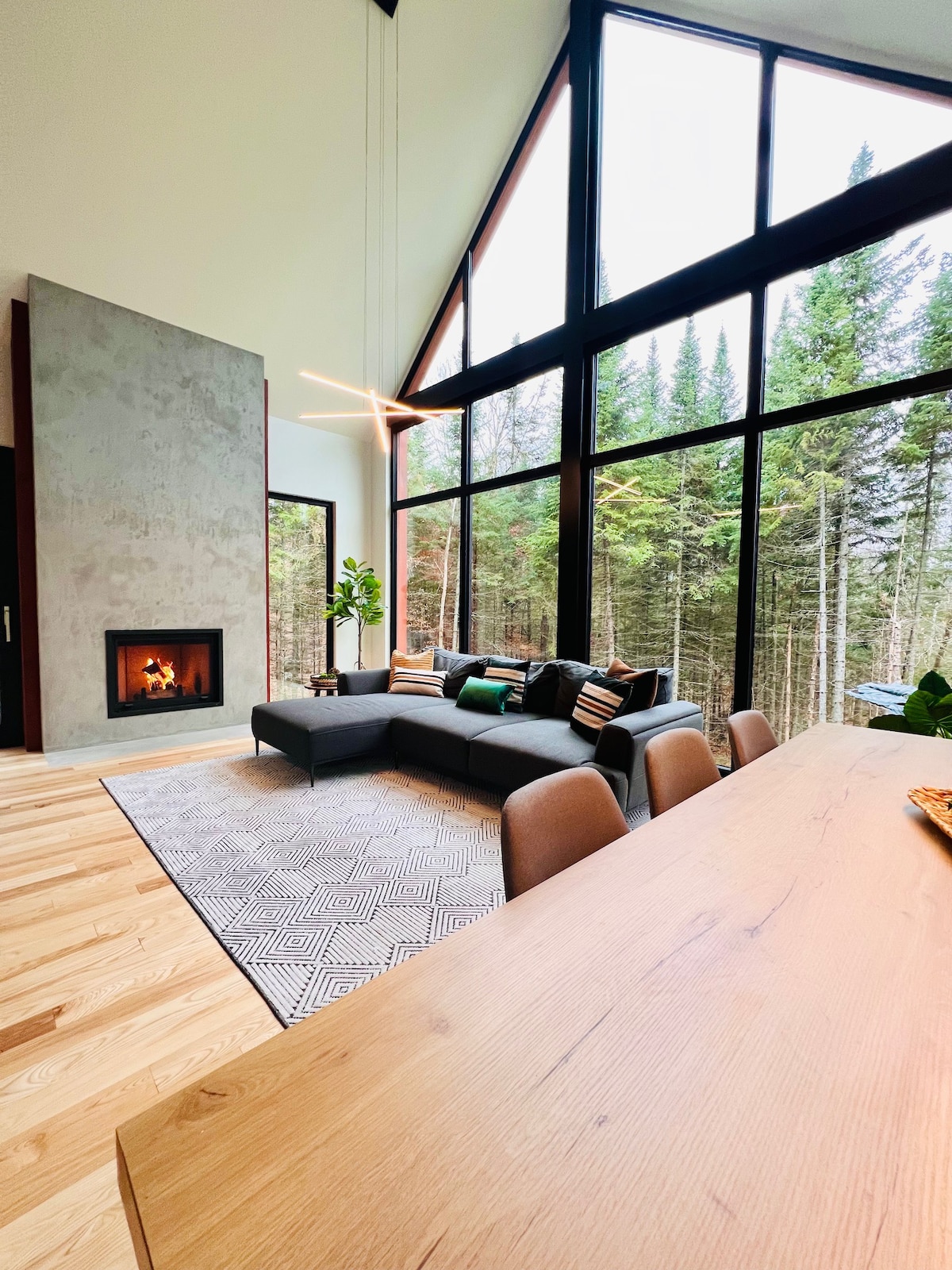 A spacious living area features a modern sectional sofa, accented with various cushions. A wall-mounted fireplace adds warmth, while large floor-to-ceiling windows showcase the surrounding pine forest. A dining table sits nearby, highlighting the open-concept design and natural light flowing into the space.
