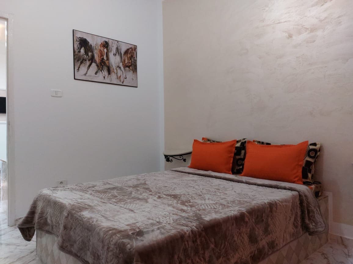 A sleeping area features a spacious bed with a light-colored blanket and two bright orange pillows. A piece of artwork displaying horses adorns the wall above. The room is finished with a smooth light-colored texture on the walls and a small side table.