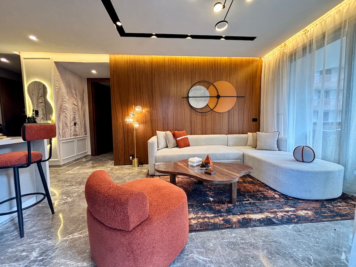 A modern living area features a curved sectional sofa in light fabric, accented by vibrant orange and brown throw pillows. A wooden coffee table sits on a patterned rug, while a stylish armchair complements the décor. Large windows allow natural light to brighten the space.