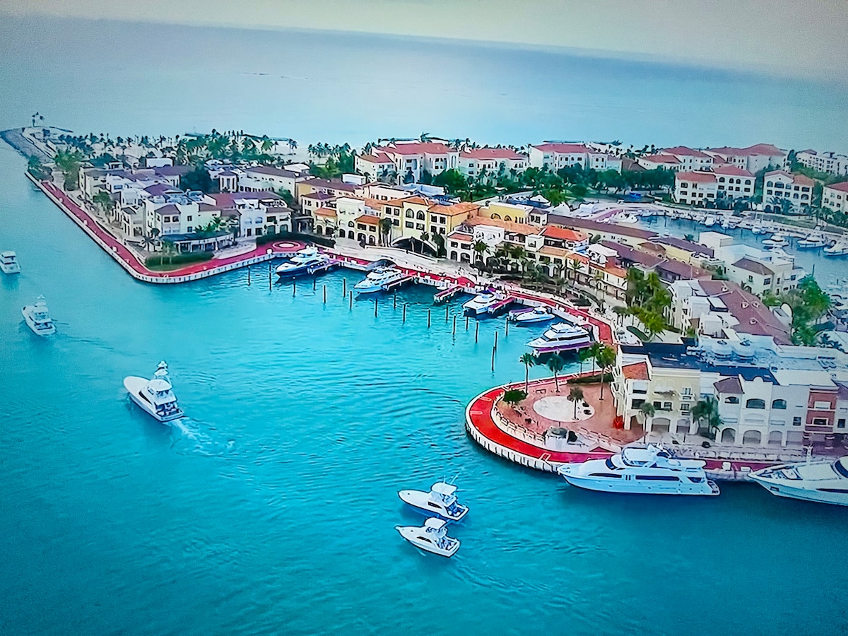 An aerial view of a vibrant marina displays numerous boats docked along the red-lined promenade. Colorful buildings surround the harbor, with lush greenery visible in the background. The clear turquoise waters reflect the coastal beauty, creating a serene and inviting atmosphere.