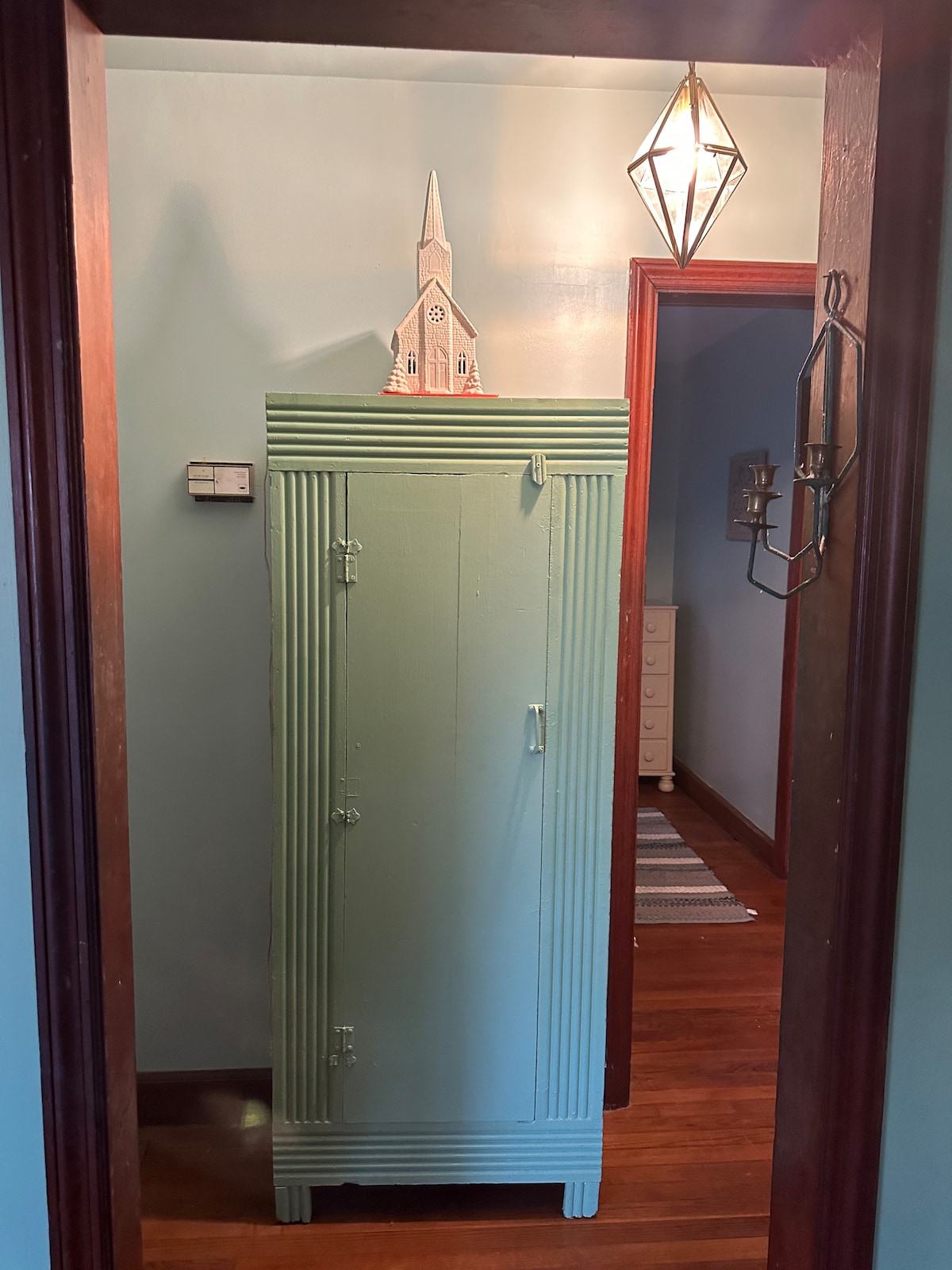 A tall, vintage-style wardrobe stands against a soft blue wall, featuring a mint green finish. Decorative elements, including a small church structure atop the wardrobe, add character. A softly lit pendant light hangs above, and a glimpse of a hallway with wooden floors can be seen in the background.