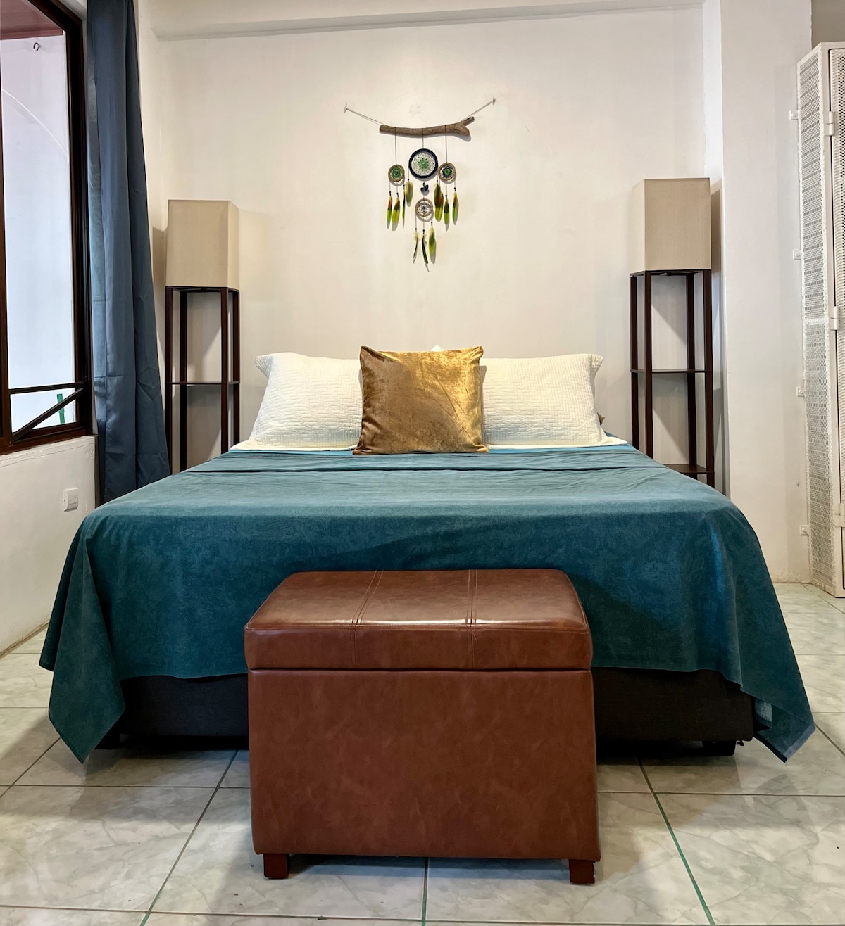 A spacious bed is dressed in a teal coverlet and accented with a golden decorative pillow. Flanking the bed are two slender, contemporary lamps atop dark stands. A small, brown ottoman sits at the foot of the bed, while natural light filters through a nearby window.