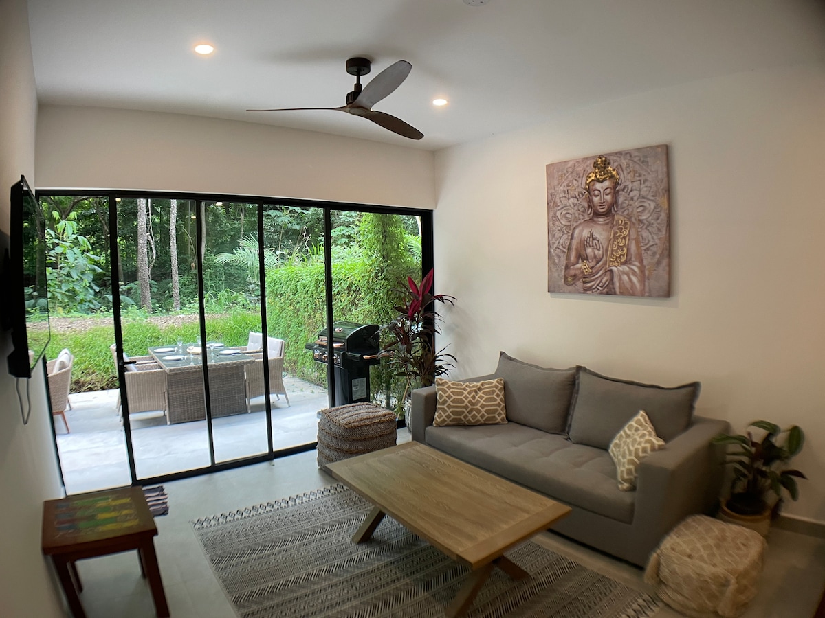 A cozy living room features a comfortable sofa and a wooden coffee table, complemented by a vibrant area rug. Large glass doors provide a view of the lush outdoor space. A wall-mounted television and ceiling fan contribute to a relaxed atmosphere.
