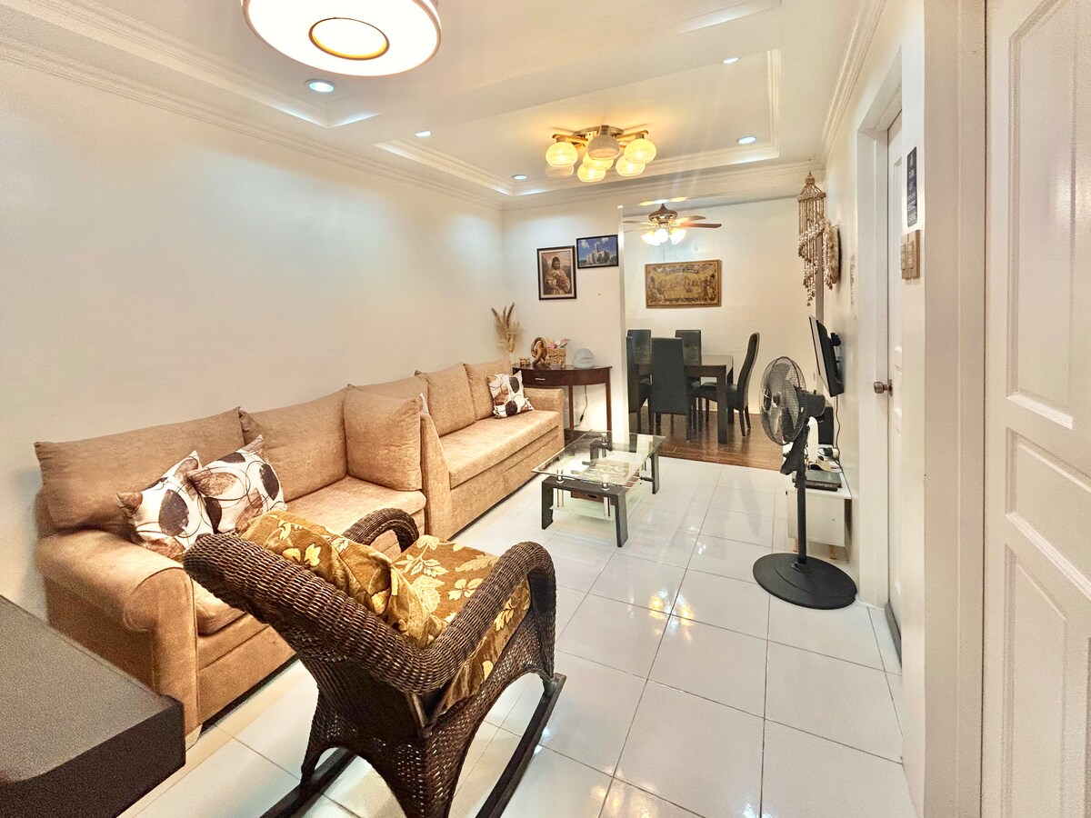 A welcoming living area features a pair of plush sofas adorned with patterned cushions, complemented by a stylish coffee table. A dark wooden dining set is visible in the background, alongside a ceiling fan and decorative wall art. Natural light opens the space, enhancing its comfort.