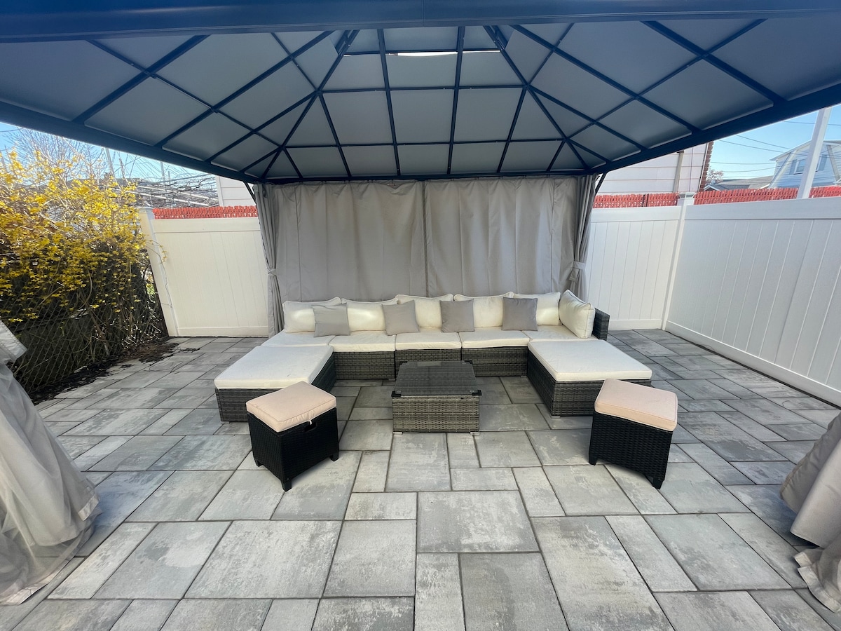 A covered outdoor seating area features a spacious arrangement of dark wicker furniture with soft white cushions. The stone patio is surrounded by a well-maintained garden, providing a private and relaxing environment. Light-gray drapes hang from the gazebo, creating a cozy ambiance.