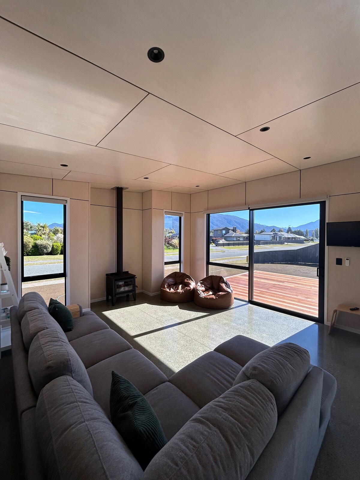 A sunlit living area is furnished with a spacious sofa facing large glass sliding doors, providing views of the outdoors. Two cozy bean bags are positioned nearby, and a minimalist wood-burning stove is installed in the corner. Light, neutral tones enhance the contemporary design.