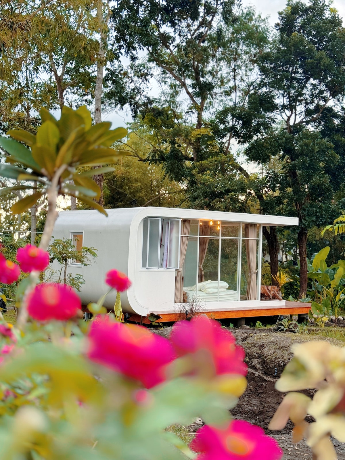 A modern cabin is surrounded by lush greenery and blooming flowers. Floor-to-ceiling glass windows provide ample natural light and offer views of the landscape. A wooden deck extends outwards, enhancing the connection to the natural surroundings.