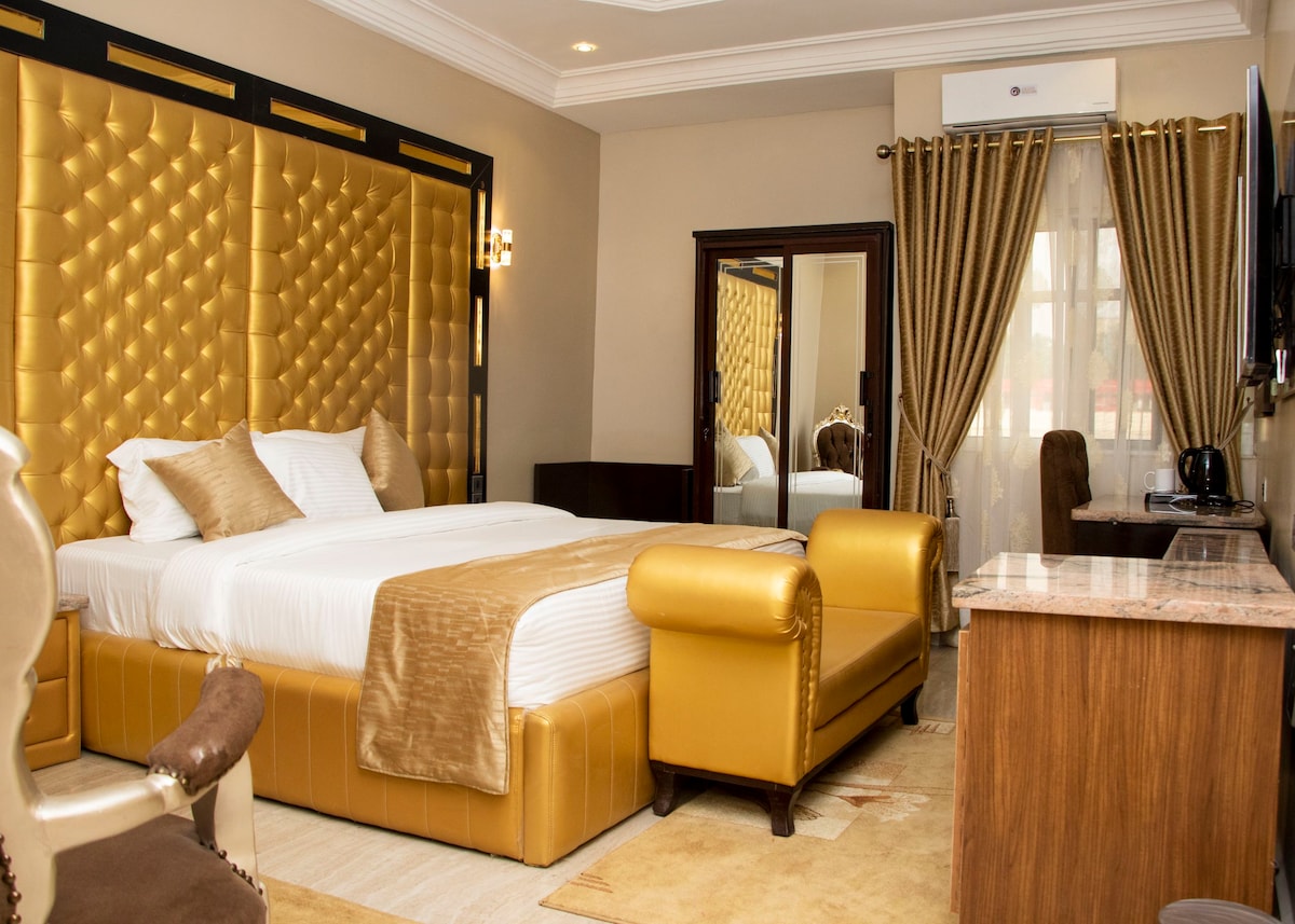 A luxurious bedroom features a large, tufted gold headboard, with a neatly made spacious bed topped with decorative pillows. A seating area with a leather lounge chair complements a stylish desk, while soft drapes frame the window, allowing natural light to fill the space.