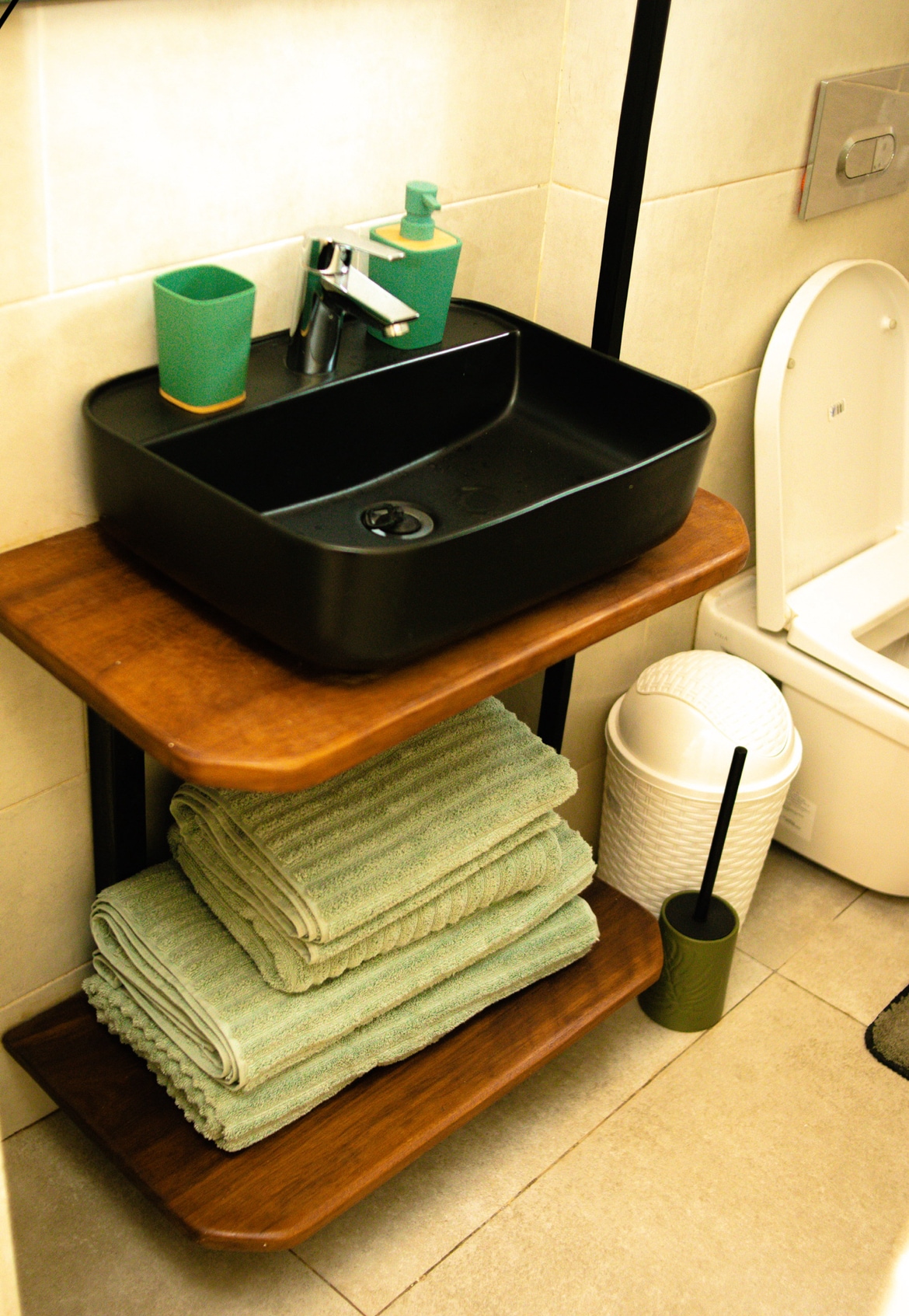 A modern bathroom sink with a sleek black basin is positioned above wooden shelves. Green towels are neatly stacked on the lower shelf, with grooming supplies and a decorative green cup placed beside the sink. A toilet is visible in the background.