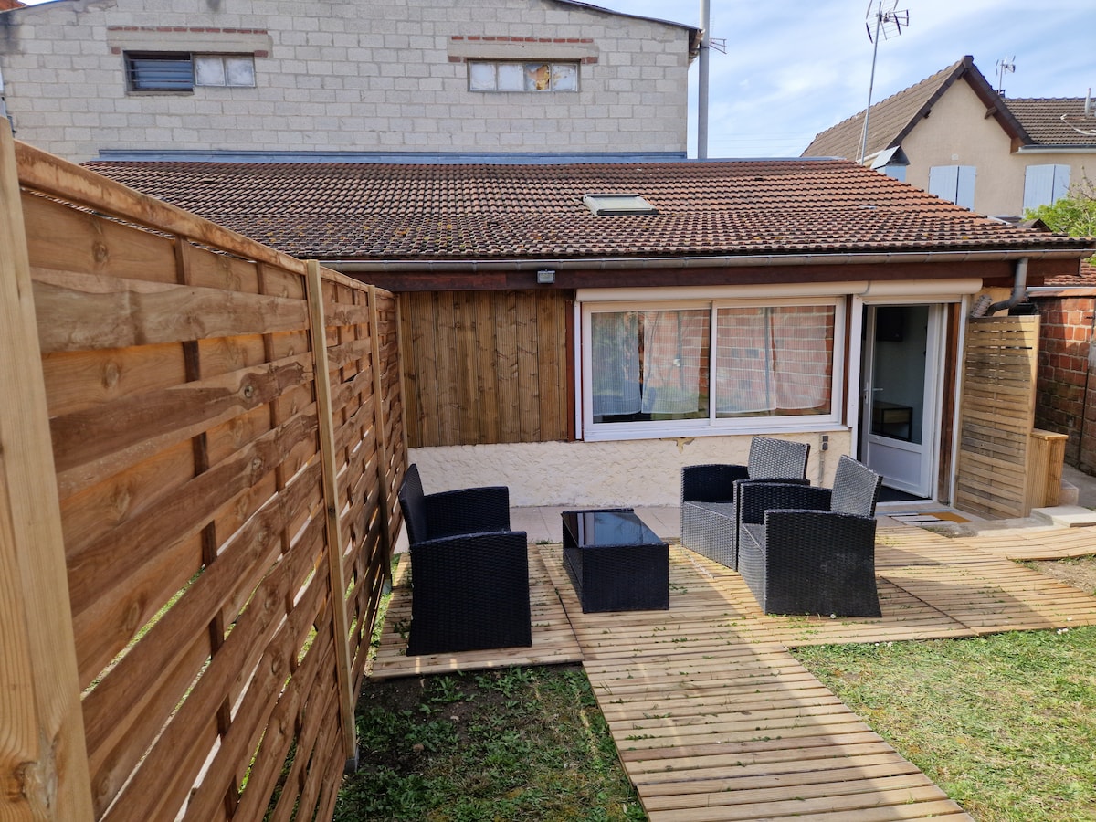 An outdoor seating area is featured, consisting of dark wicker chairs surrounding a glass-top table. The wood deck leads to a door that opens into the interior space. A wooden fence provides privacy, and a grassy area is present beside the deck.