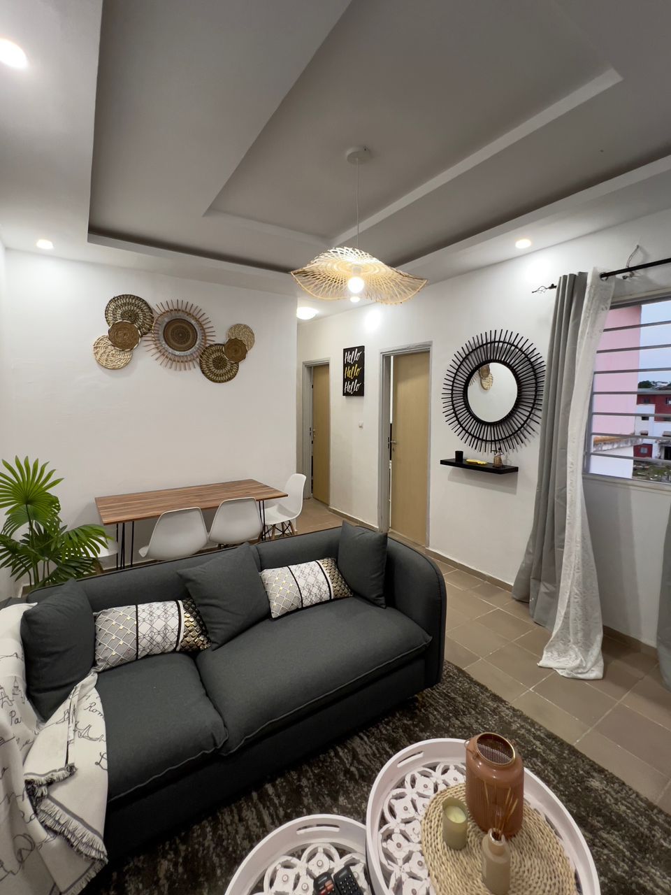 The living area features a modern gray sofa adorned with decorative cushions. A round dining table with white chairs sits in the background. Wall decor includes woven baskets and a sunburst mirror. Natural light enters through a window, complemented by light curtains.